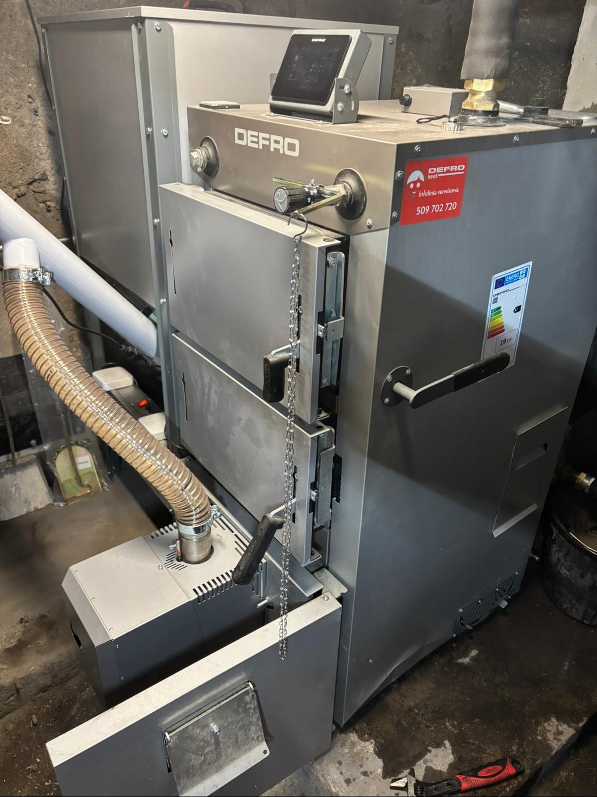 Szary, nowoczesny piec Defro z panelem sterowania i łańcuchem, widoczny w kotłowni. Na obudowie naklejka z numerem infolinii serwisowej.