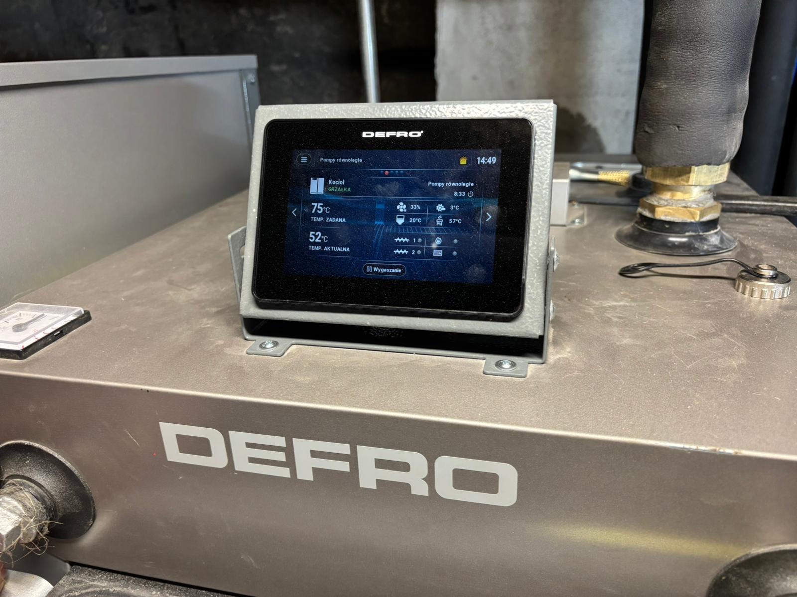 Panel sterowania kotła DEFRO z wyświetlanymi parametrami pracy, widoczne rury instalacji. Precyzyjny monitoring i kontrola systemu grzewczego.