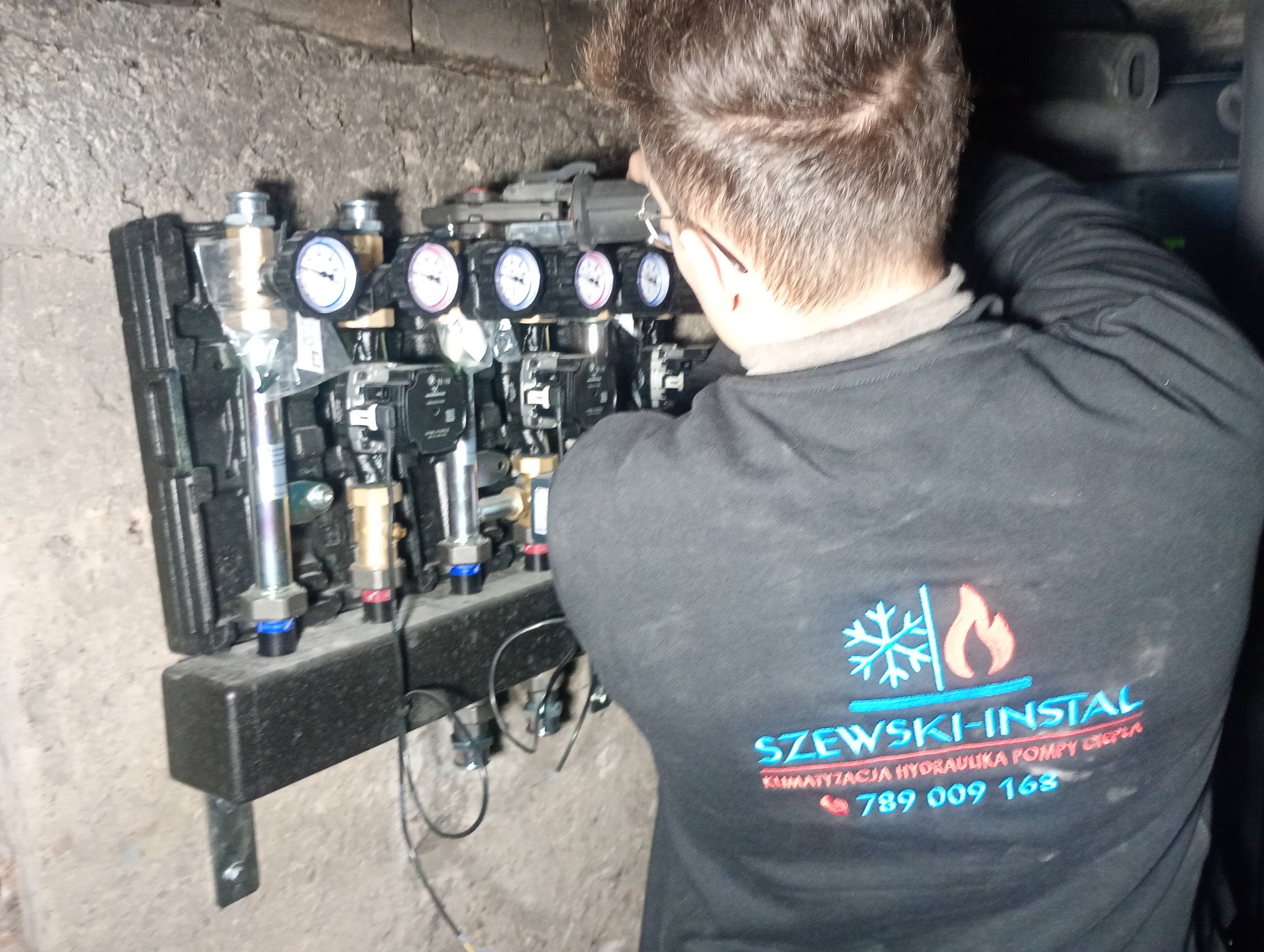 Instalator w trakcie montażu rozdzielacza ogrzewania podłogowego z manometrami, widoczny fragment ściany i logo firmy na bluzie montera.