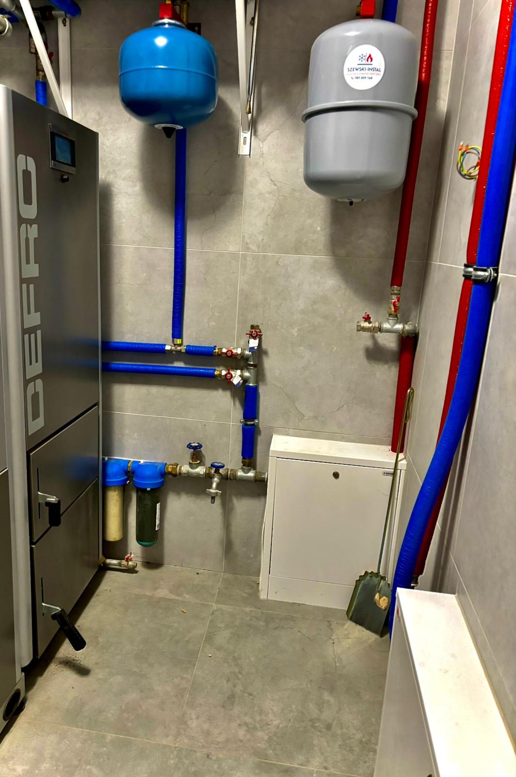 Kotłownia z nowoczesnym piecem DEfro, zbiornikami wyrównawczymi i filtrami wody. Instalacja z rur w kolorach niebieskim i czerwonym. Szare płytki na ścianach i podłodze.