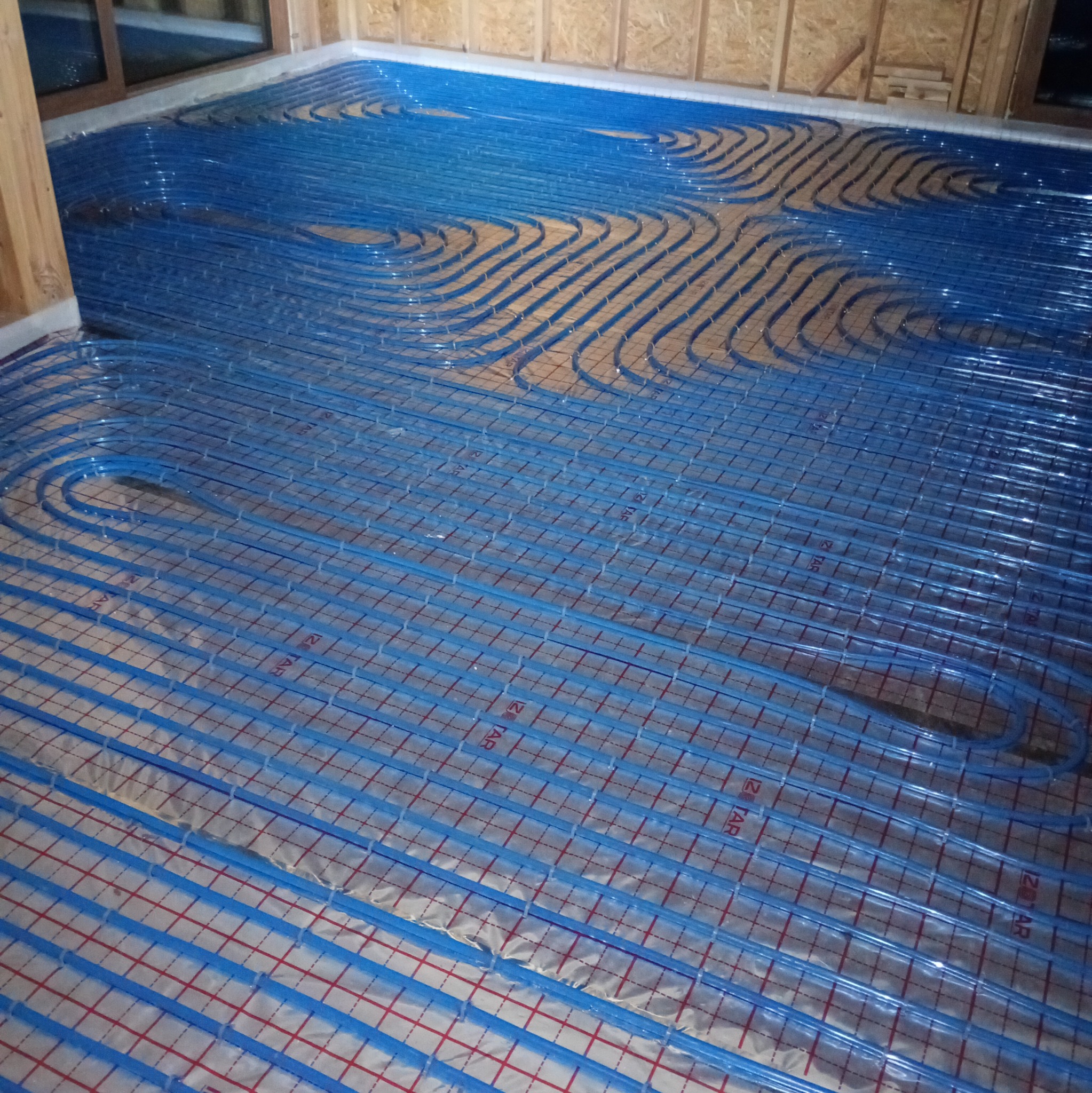 Niebieskie rury ogrzewania podłogowego ułożone na folii z siatką, widoczne drewniane elementy konstrukcji budynku. Układ spiralny rur.