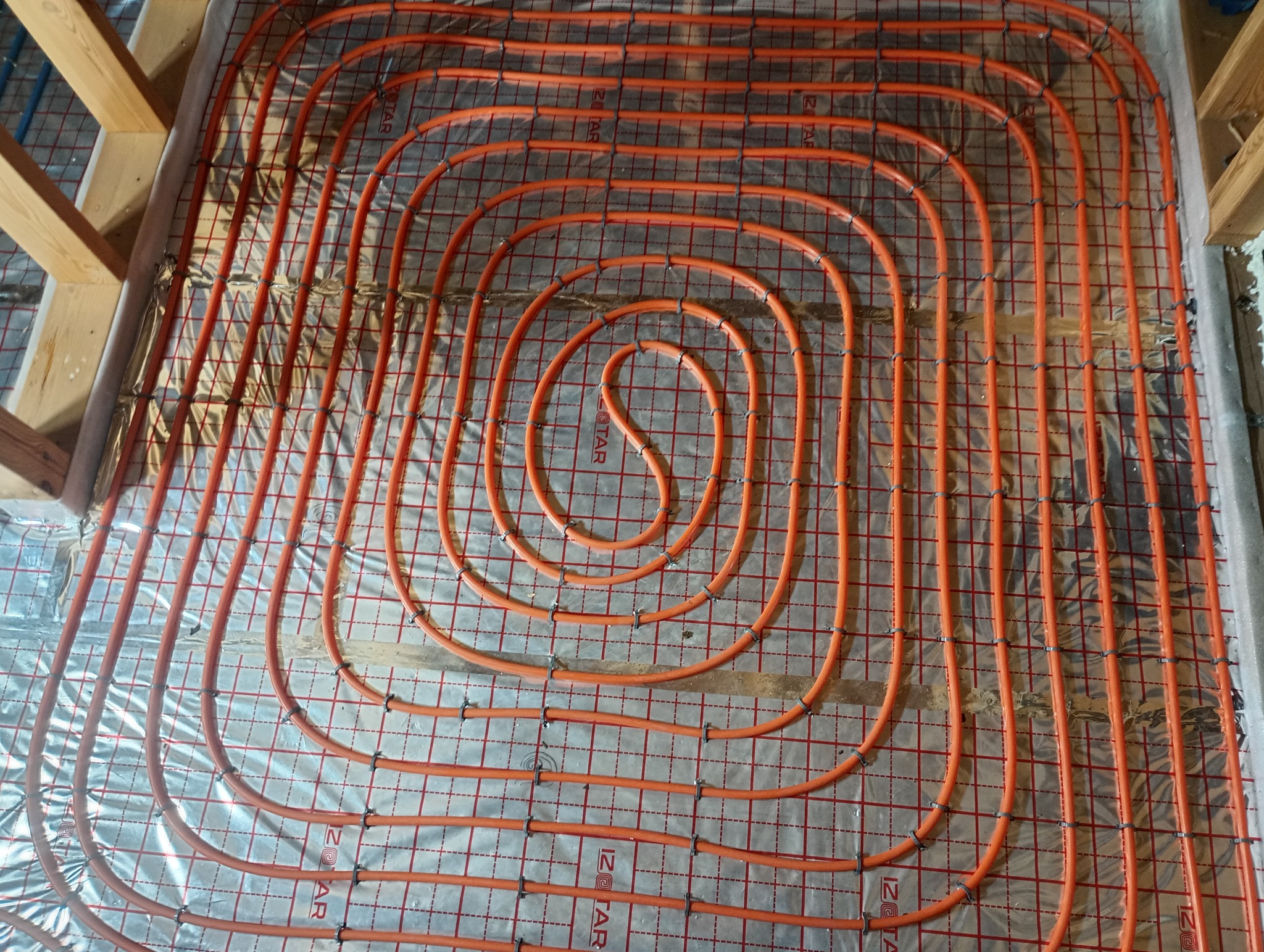 Pomarańczowe rury ogrzewania podłogowego ułożone spiralnie na folii izolacyjnej z siatką, przymocowane plastikowymi uchwytami. Widoczne drewniane elementy konstrukcyjne.