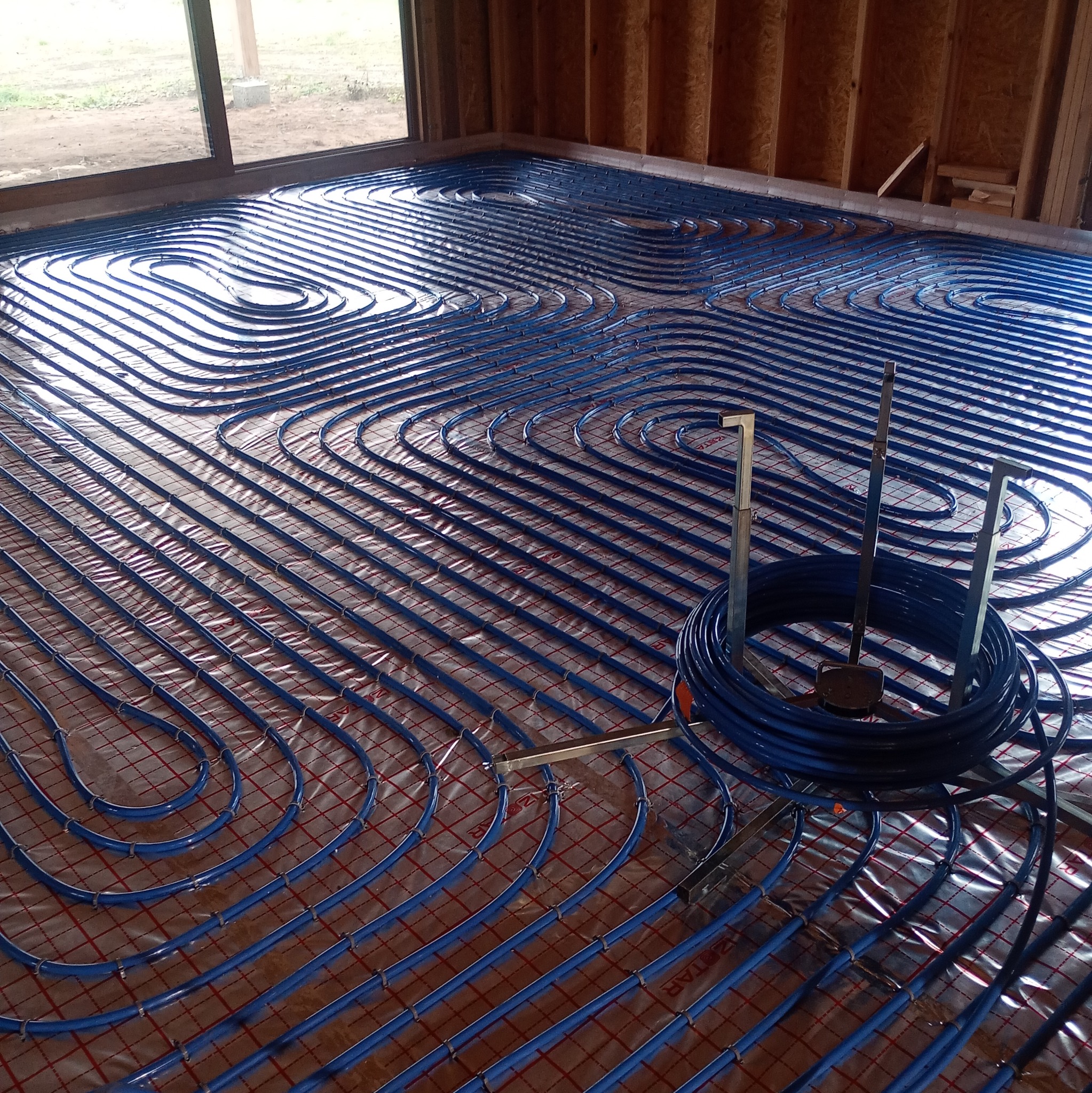 Niebieskie rury ogrzewania podłogowego ułożone na folii z siatką, widoczne narzędzia montażowe i fragment ściany z desek. Instalacja w trakcie realizacji.