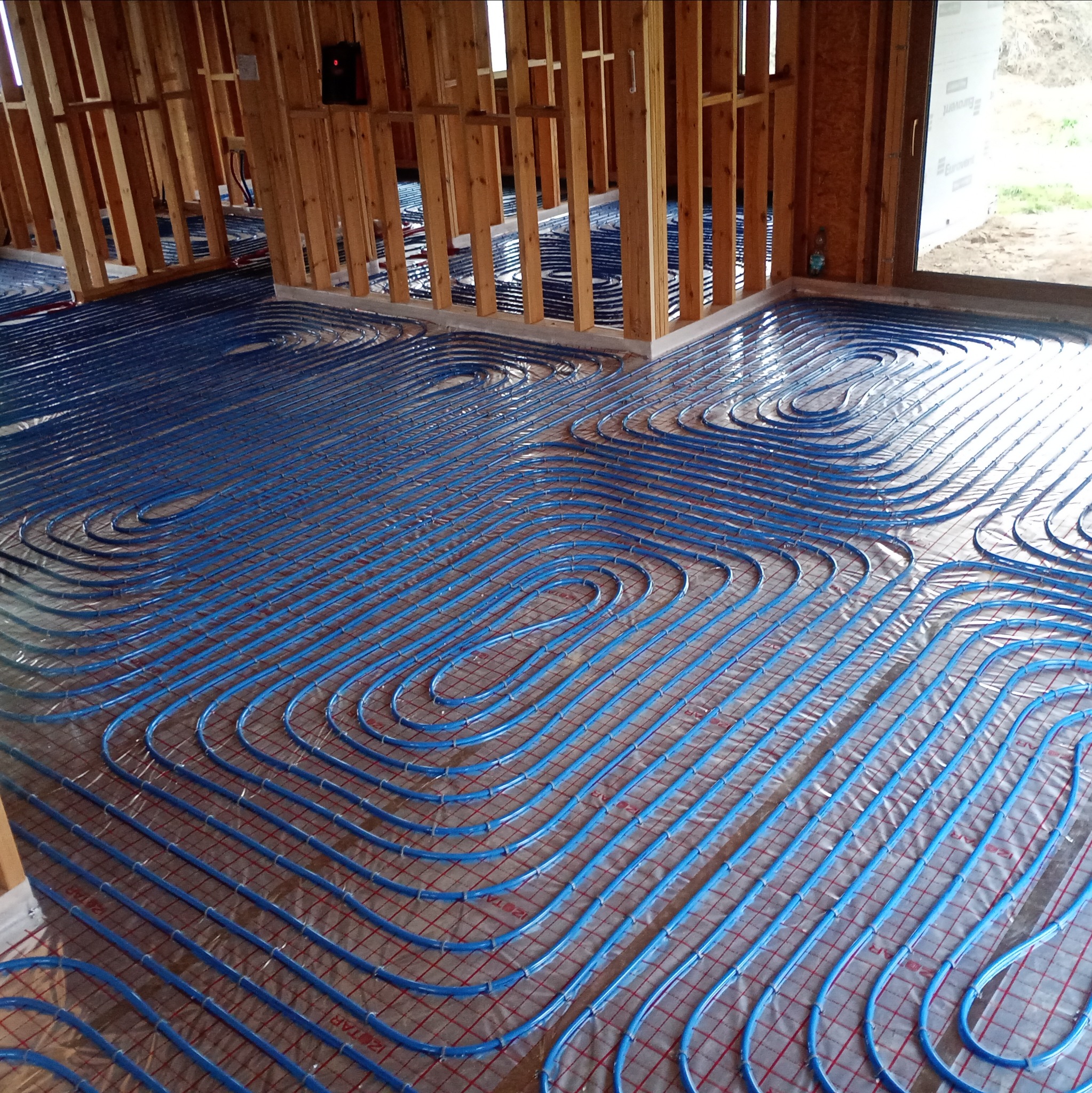 Niebieskie rury ogrzewania podłogowego ułożone na folii odblaskowej i siatce w surowym wnętrzu domu w trakcie budowy. Widoczne drewniane ramy ścian.