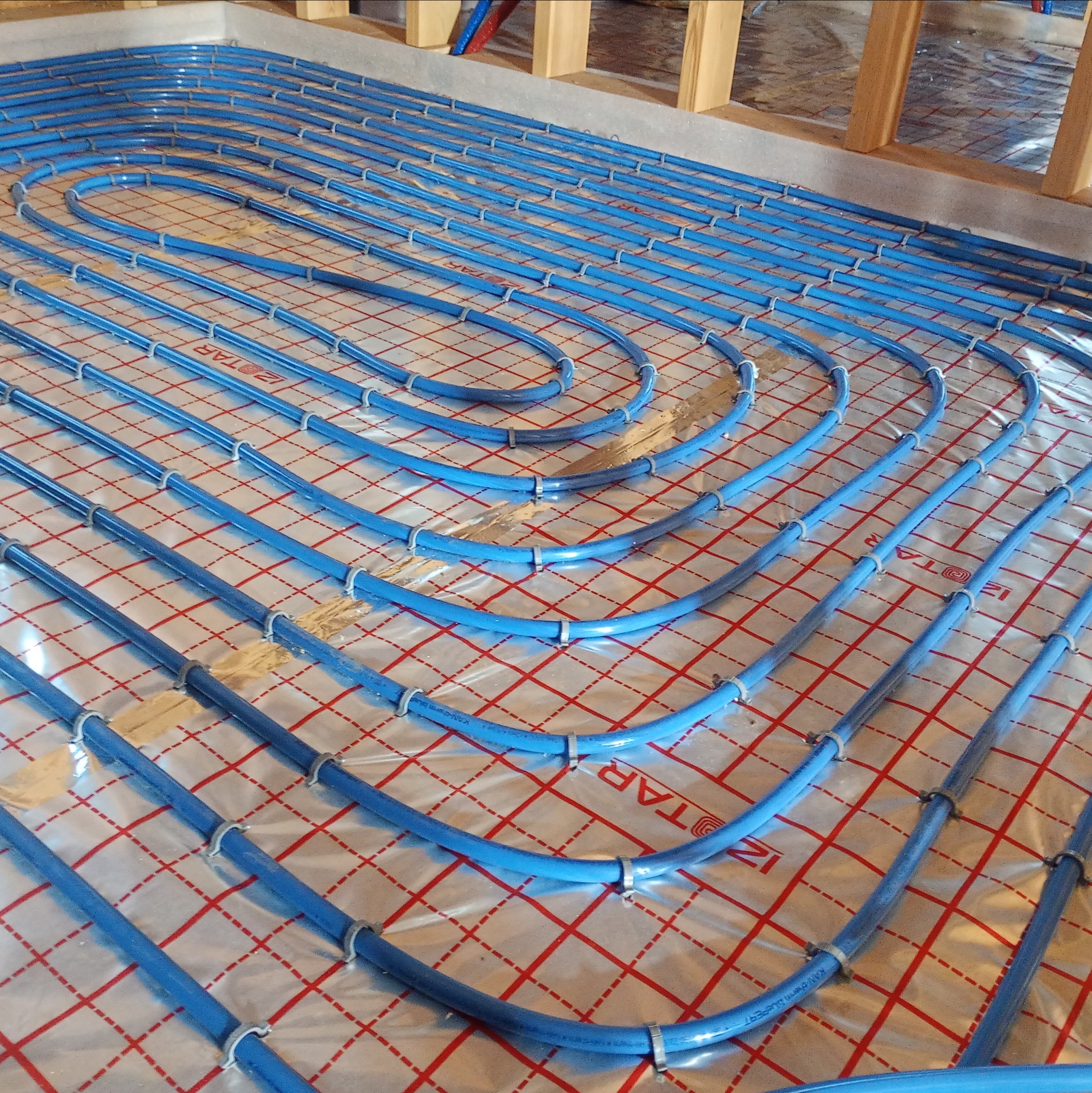 Niebieskie rury ogrzewania podłogowego ułożone na folii z czerwoną kratką, przymocowane plastikowymi uchwytami. Widoczne drewniane elementy konstrukcyjne.