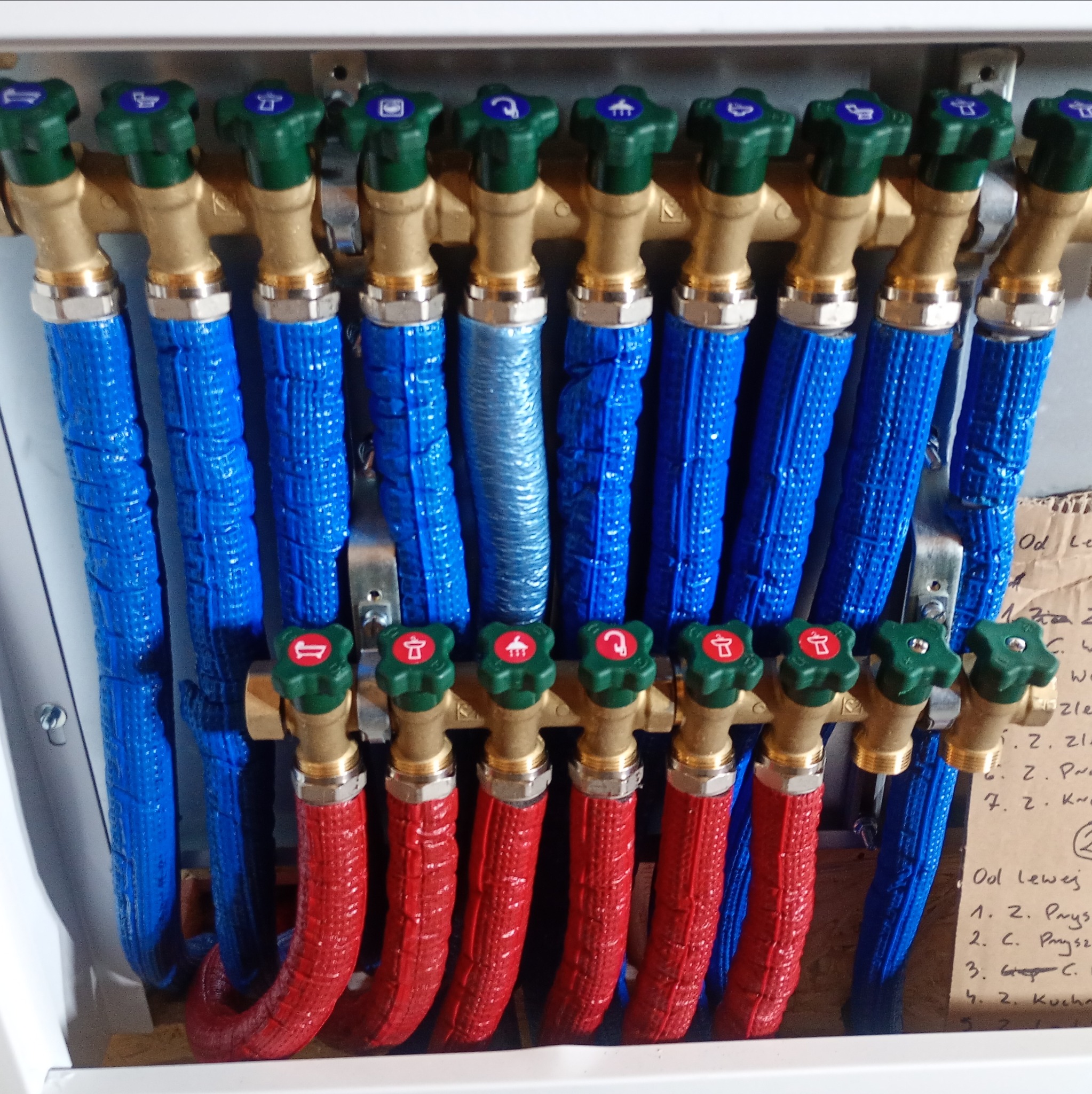 Rozdzielacz instalacji wodnej z rurami w izolacji: czerwone (ciepła woda), niebieskie (zimna). Zielone zawory z ikonami. Widoczne notatki na kartonie.