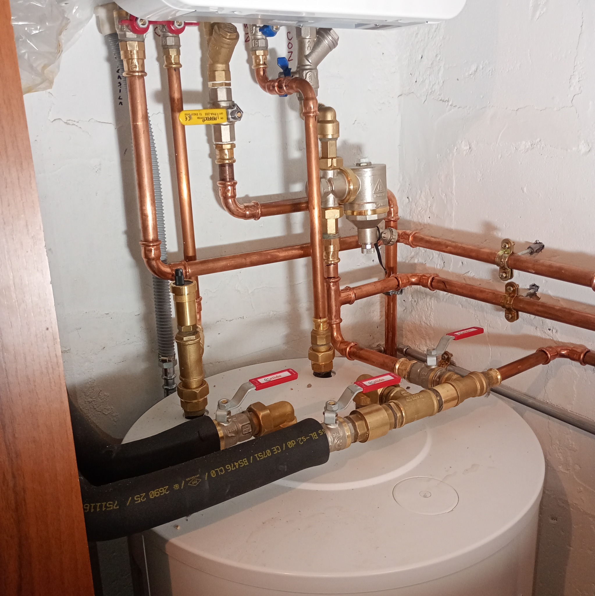 Instalacja hydrauliczna z miedzianymi rurami i zaworami Calido, zamontowana na białym zbiorniku w pomieszczeniu z białą, nierówną ścianą. Widoczne złącza i elementy mocujące.