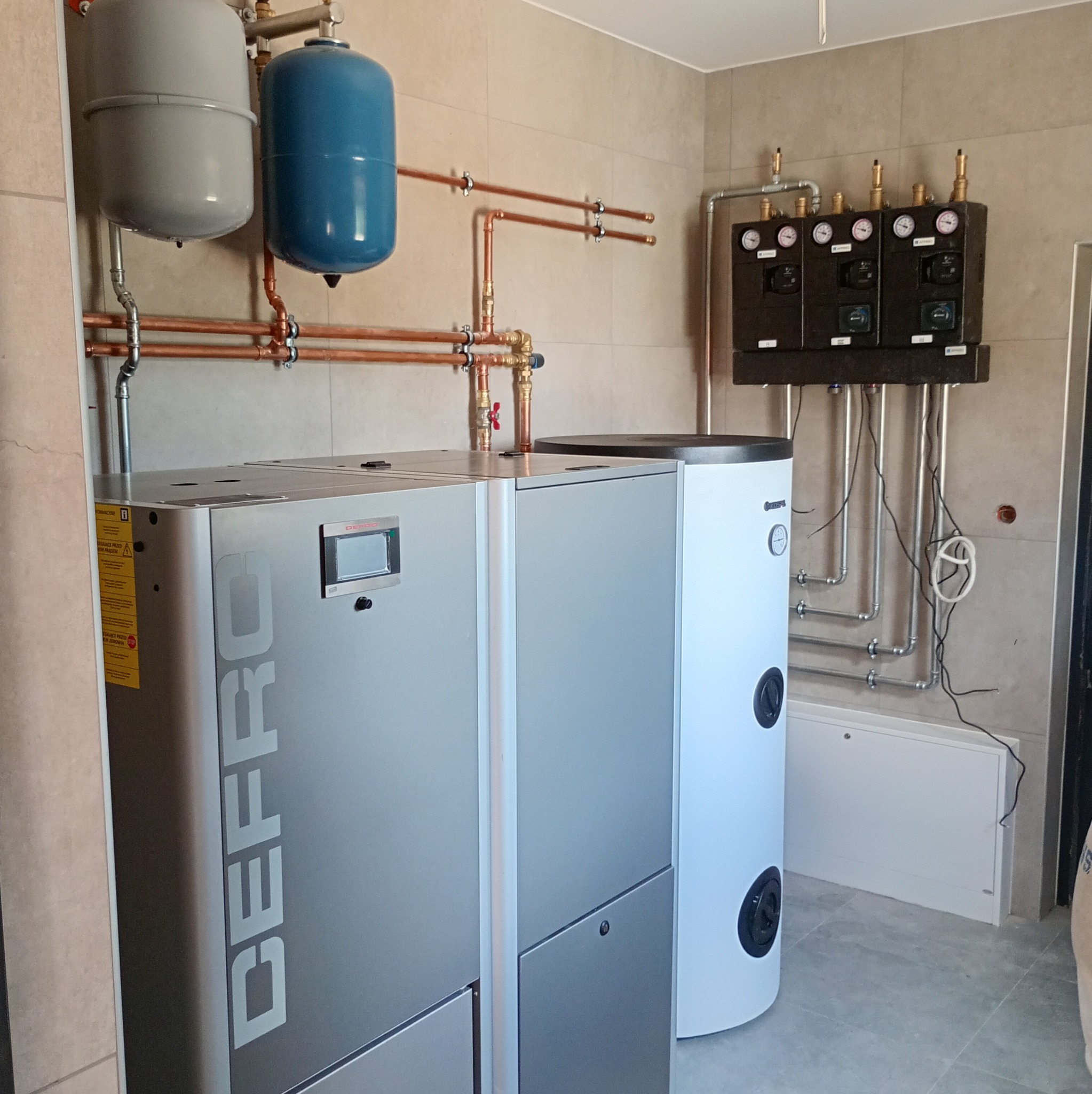 Nowoczesna kotłownia z piecem Defro, zbiornikiem buforowym i rozbudowaną instalacją hydrauliczną z miedzianymi rurami i czarnym panelem sterowania.