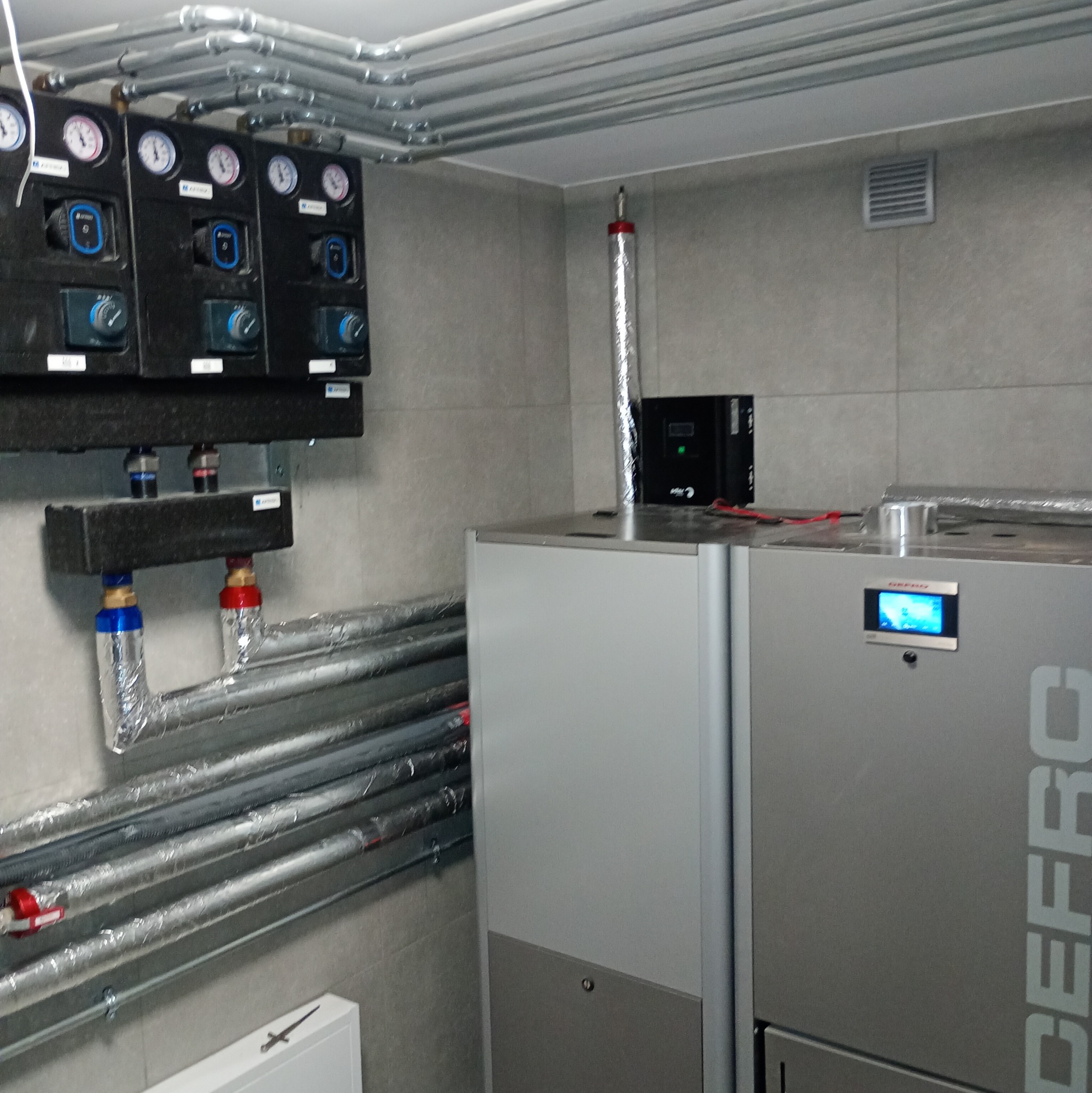 Złożona instalacja grzewcza z rurami w izolacji, czarnymi panelami kontrolnymi z manometrami i srebrnym piecem z wyświetlaczem w pomieszczeniu z szarą płytką.