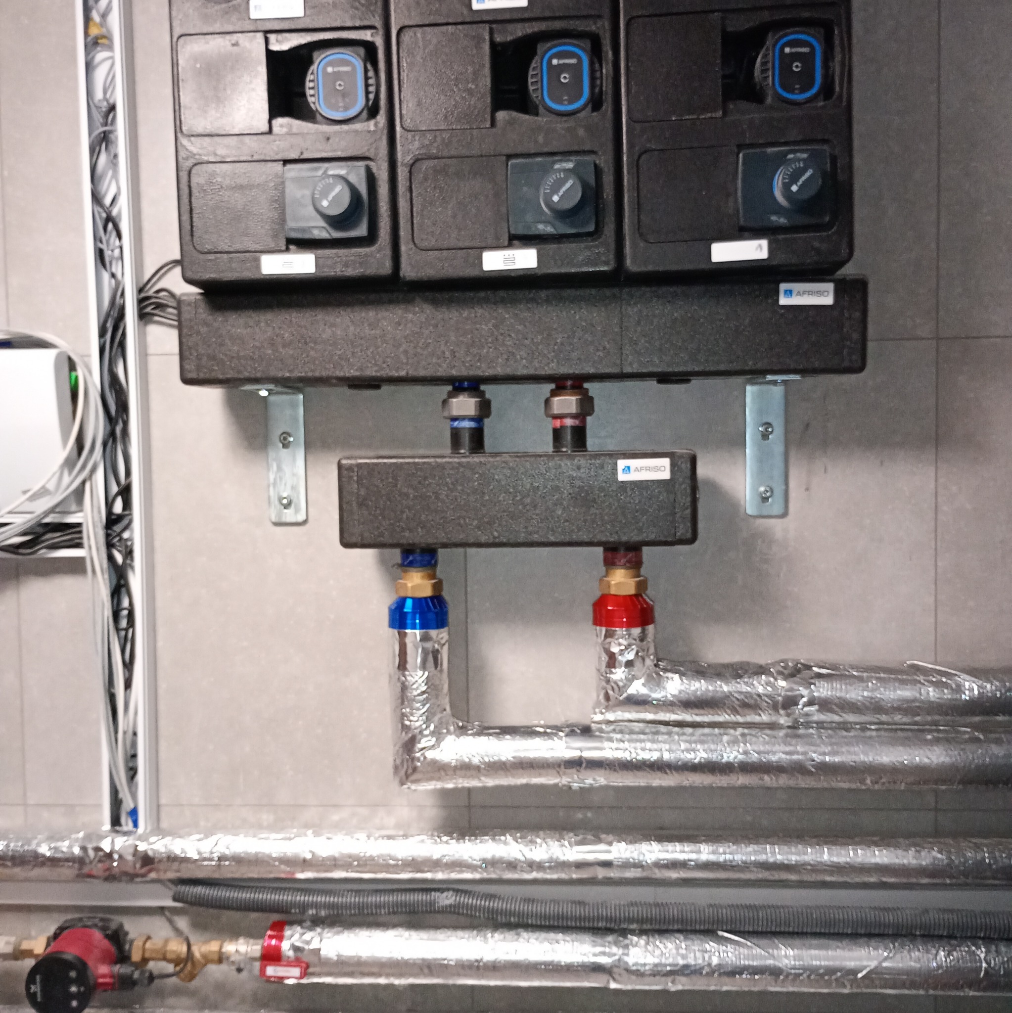 Rozdzielacz ogrzewania podłogowego z pompami obiegowymi, rury w izolacji aluminiowej, zamontowany na szarej ścianie. Widoczne elementy sterujące i złączki.