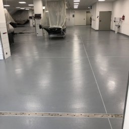 Piotr Biegun - Szara, gładka posadzka w sterylnym pomieszczeniu z odpływem liniowym. Sprzęt medyczny przykryty folią ochronną. Białe ściany i sufit z oświetleniem rastrowym.