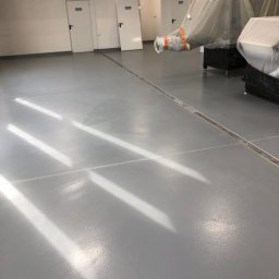 Piotr Biegun - Jasnoszara, gładka posadzka w hali przemysłowej z widocznymi dylatacjami i refleksami światła. W tle białe drzwi i sprzęt zabezpieczony folią malarską.