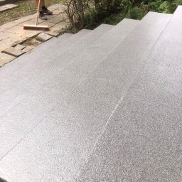 Piotr Biegun - Schody z szarego mikrocementu, czyszczone szczotką przez pracownika w rękawicach. W tle zieleń i fragment samochodu. Wykończenie zewnętrzne.