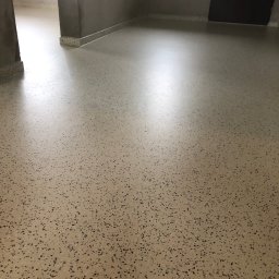 Piotr Biegun - Beżowa posadzka z ciemnymi drobinkami w jasnym pomieszczeniu z szarą ścianą i ciemnymi drzwiami. Widoczny fragment listwy przypodłogowej w tym samym stylu.