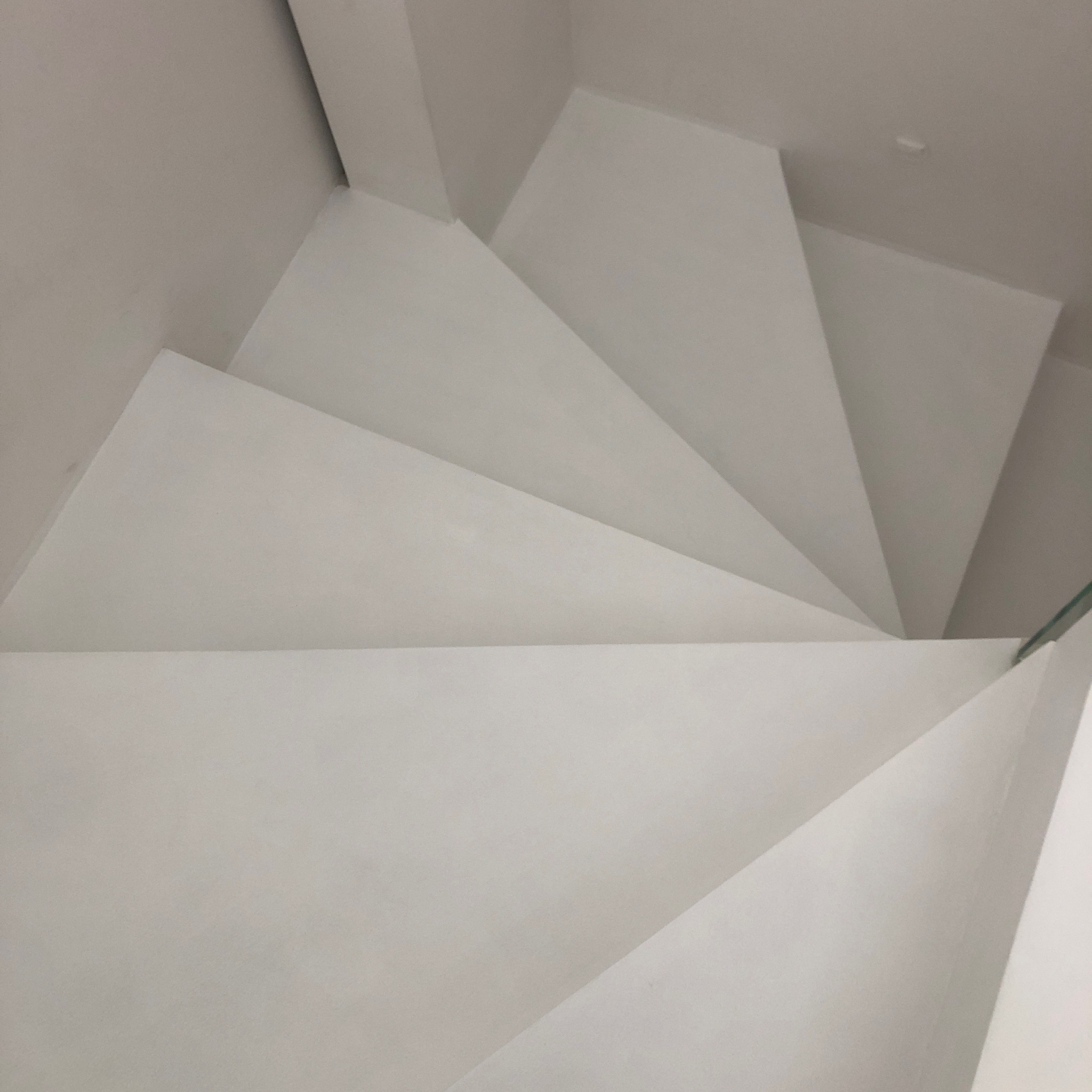 Minimalistyczne, białe schody z gładką powierzchnią, widziane z góry, z delikatnym oświetleniem podkreślającym geometrię stopni. Widoczny fragment szklanej balustrady.