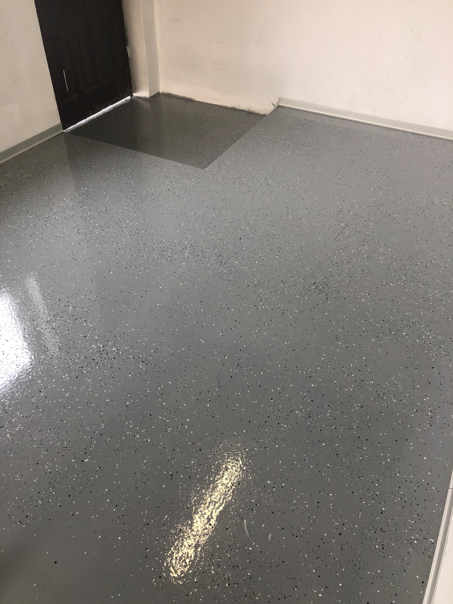 Szara, błyszcząca posadzka z drobnymi, ciemnymi i jasnymi plamkami, widoczna przy białej listwie przypodłogowej i ciemnych drzwiach. Nowoczesne wykończenie podłogi.