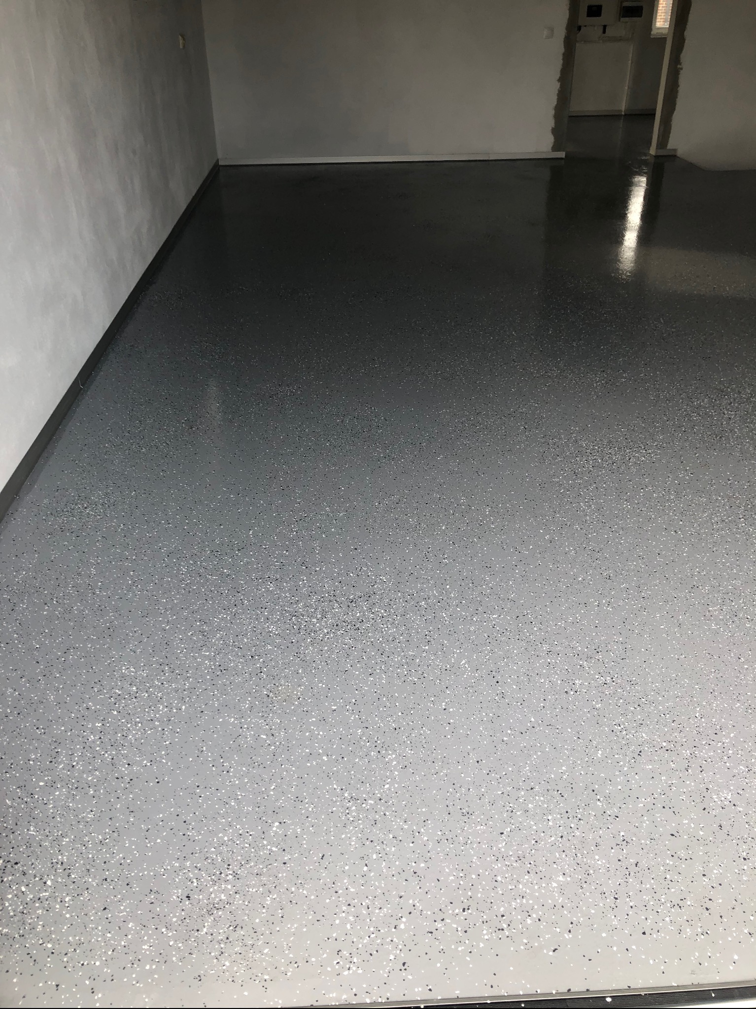 Błyszcząca, szara posadzka żywiczna z drobnymi, ciemnymi i jasnymi plamkami, odbijająca światło w pustym pomieszczeniu z szarymi ścianami i widocznym wejściem.