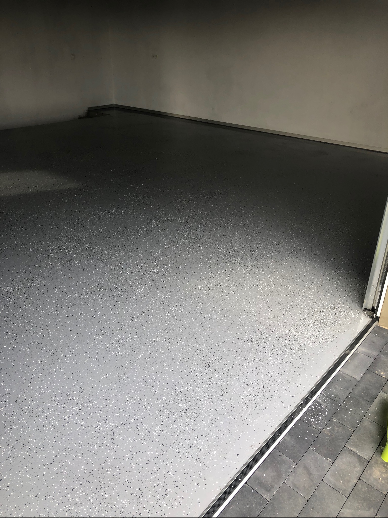 Szara posadzka z drobnymi, kolorowymi wtrąceniami, wykonana w garażu. Widoczny fragment progu i kostki brukowej na zewnątrz. Oświetlenie górne, jednolite.