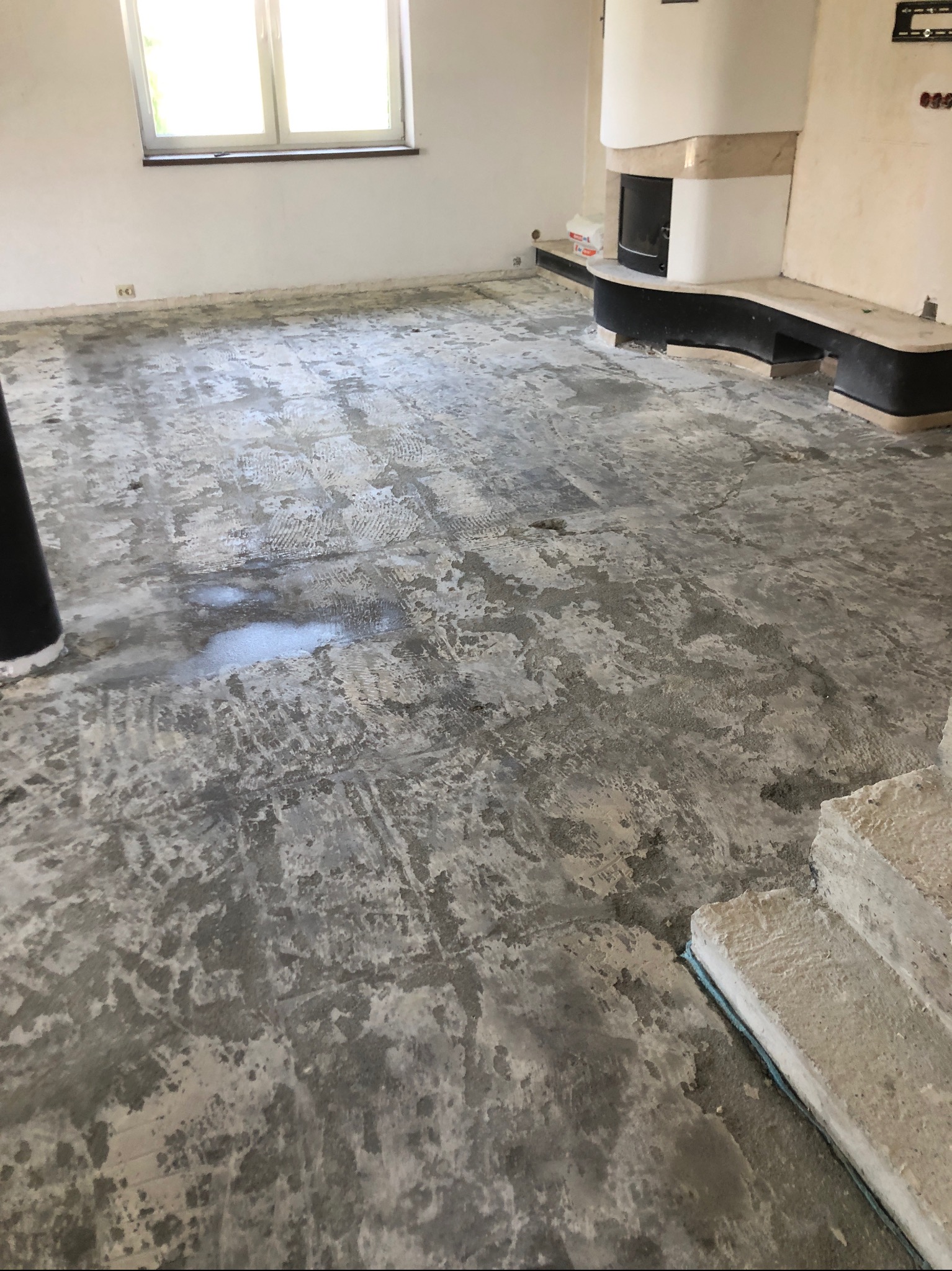 Przygotowanie podłoża pod posadzkę w jasnym wnętrzu z kominkiem. Widoczne ślady po szlifowaniu i pozostałości starej posadzki. Okno w tle.