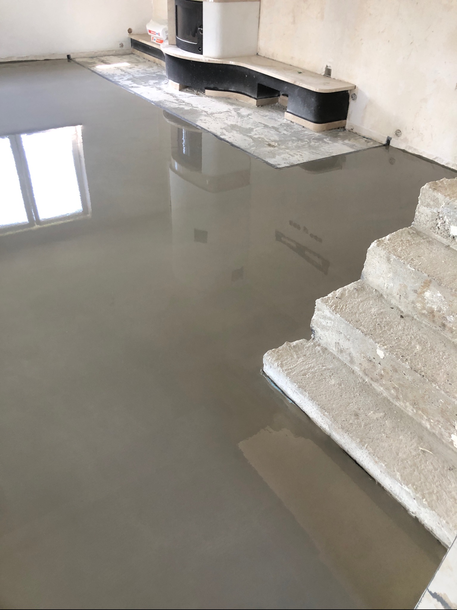 Świeżo wylana, gładka, szara posadzka w pomieszczeniu z oknem i schodami. Widoczne ślady po narzędziach i refleksy światła na mokrej powierzchni.