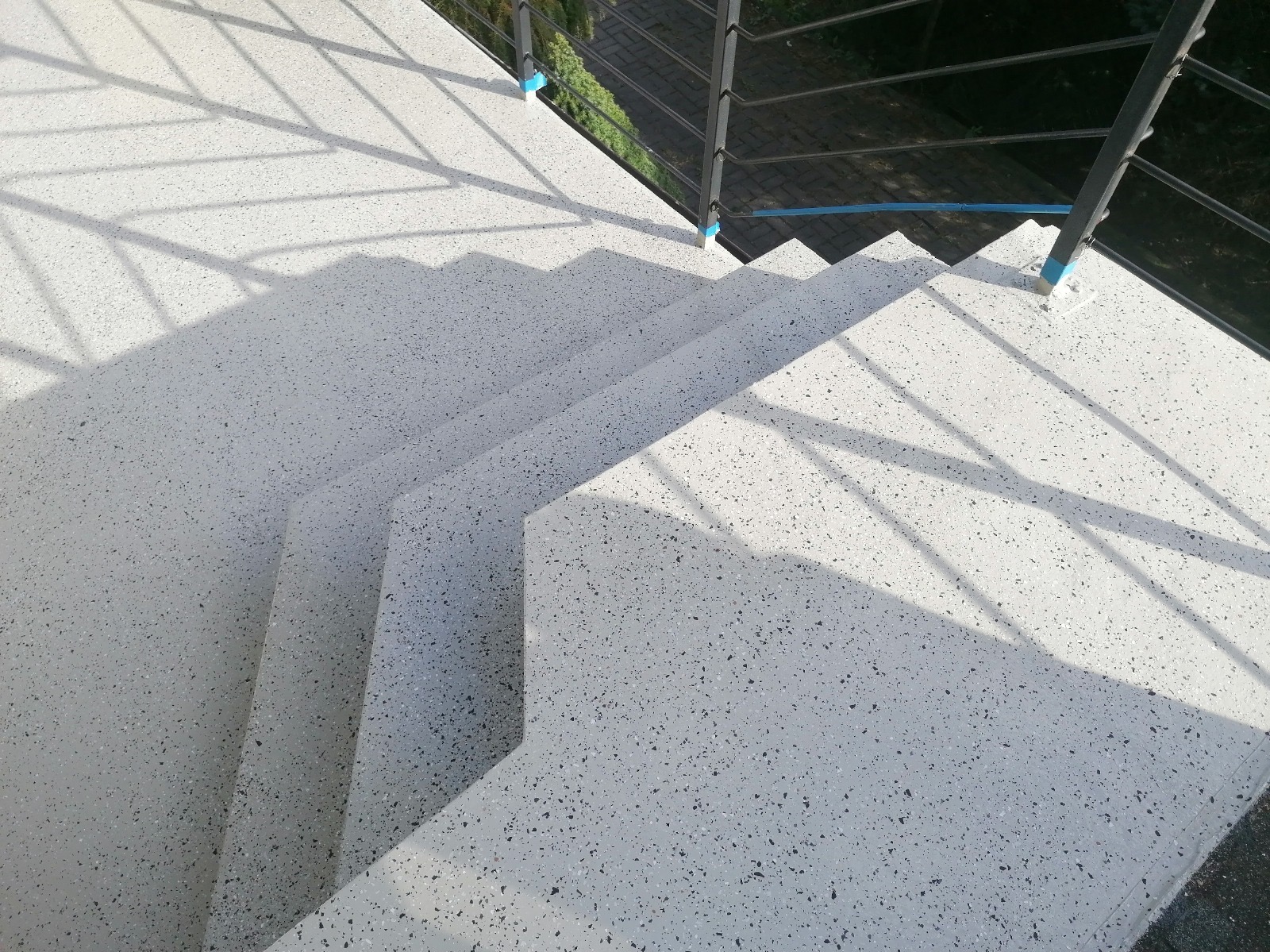 Schody z jasnoszarej posadzki lastryko z czarnymi drobinkami, oświetlone naturalnym światłem, z widoczną metalową barierką i cieniem.