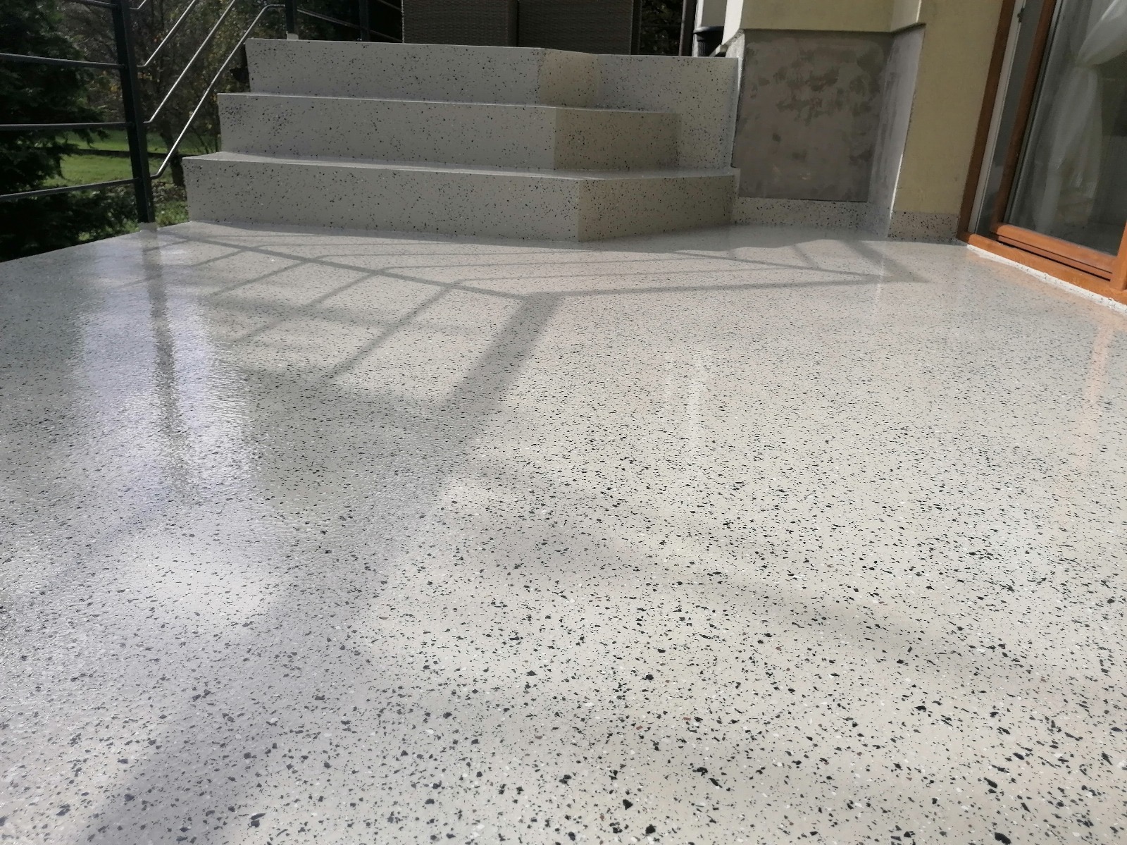 Jasnoszara, błyszcząca posadzka z drobnymi, ciemnymi plamkami na balkonie, prowadząca do schodów. Widoczne refleksy światła i cień barierki.