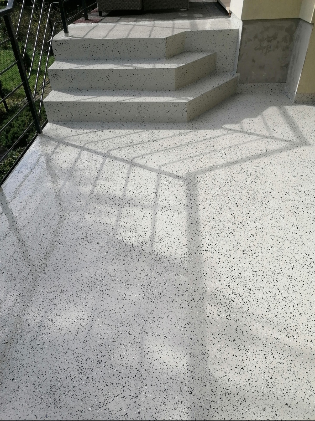 Biała posadzka żywiczna z czarnymi drobinkami na balkonie, schody z tego samego materiału, cień barierki. Nowoczesne wykończenie balkonu.