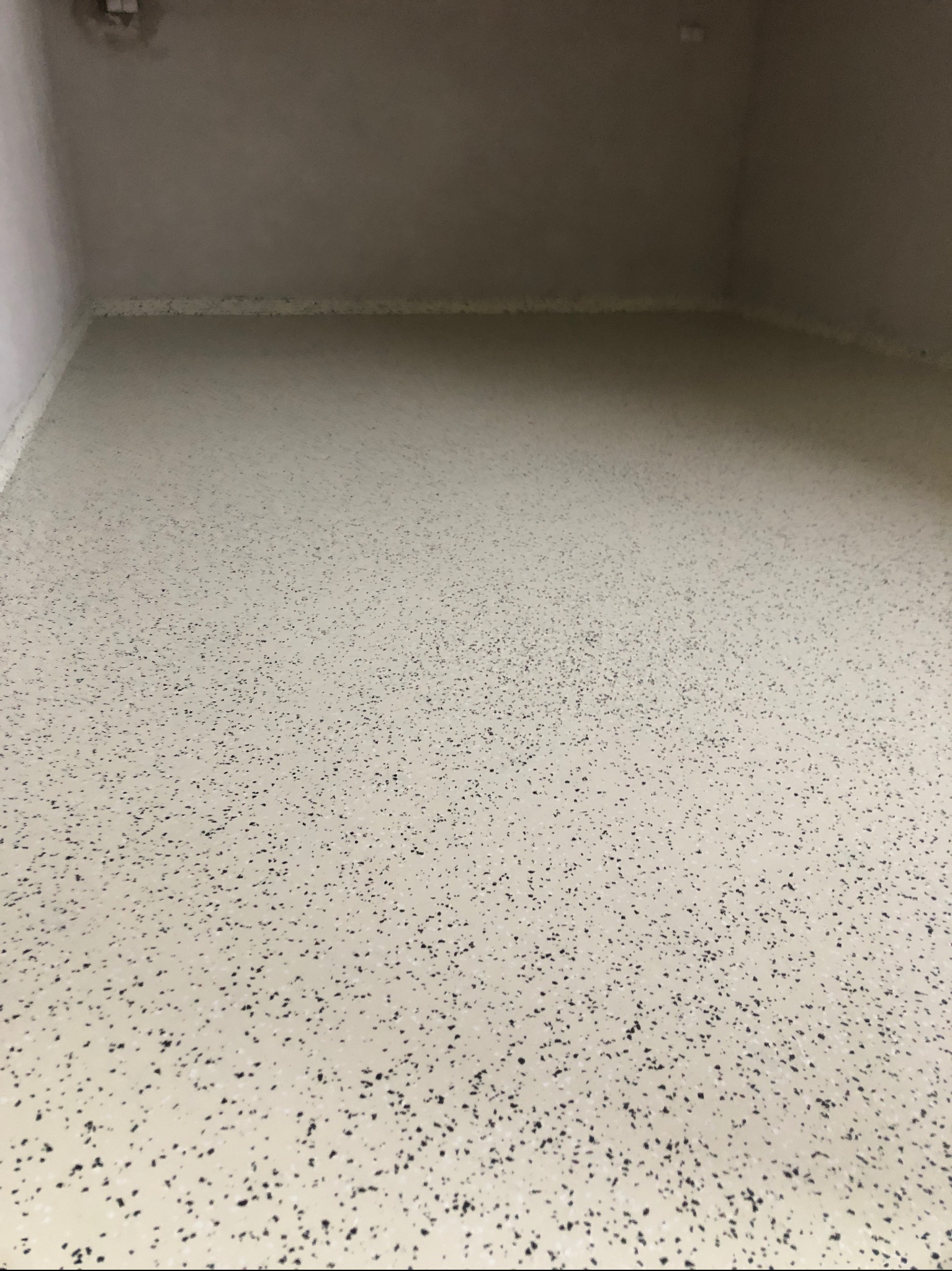 Jasna, gładka posadzka z ciemnymi, nieregularnymi plamkami, wypełniająca całą przestrzeń kadru, w rogu widoczny fragment ściany i sufitu.