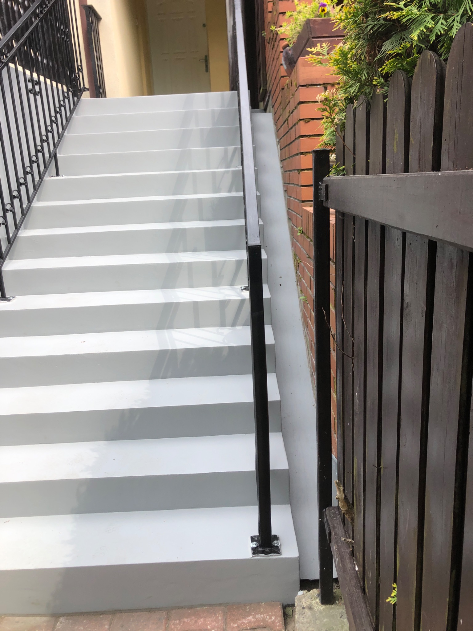 Odnowione schody zewnętrzne z szarą, gładką powierzchnią i czarną, metalową barierką. Widoczny fragment ceglanego muru i drewnianego płotu.