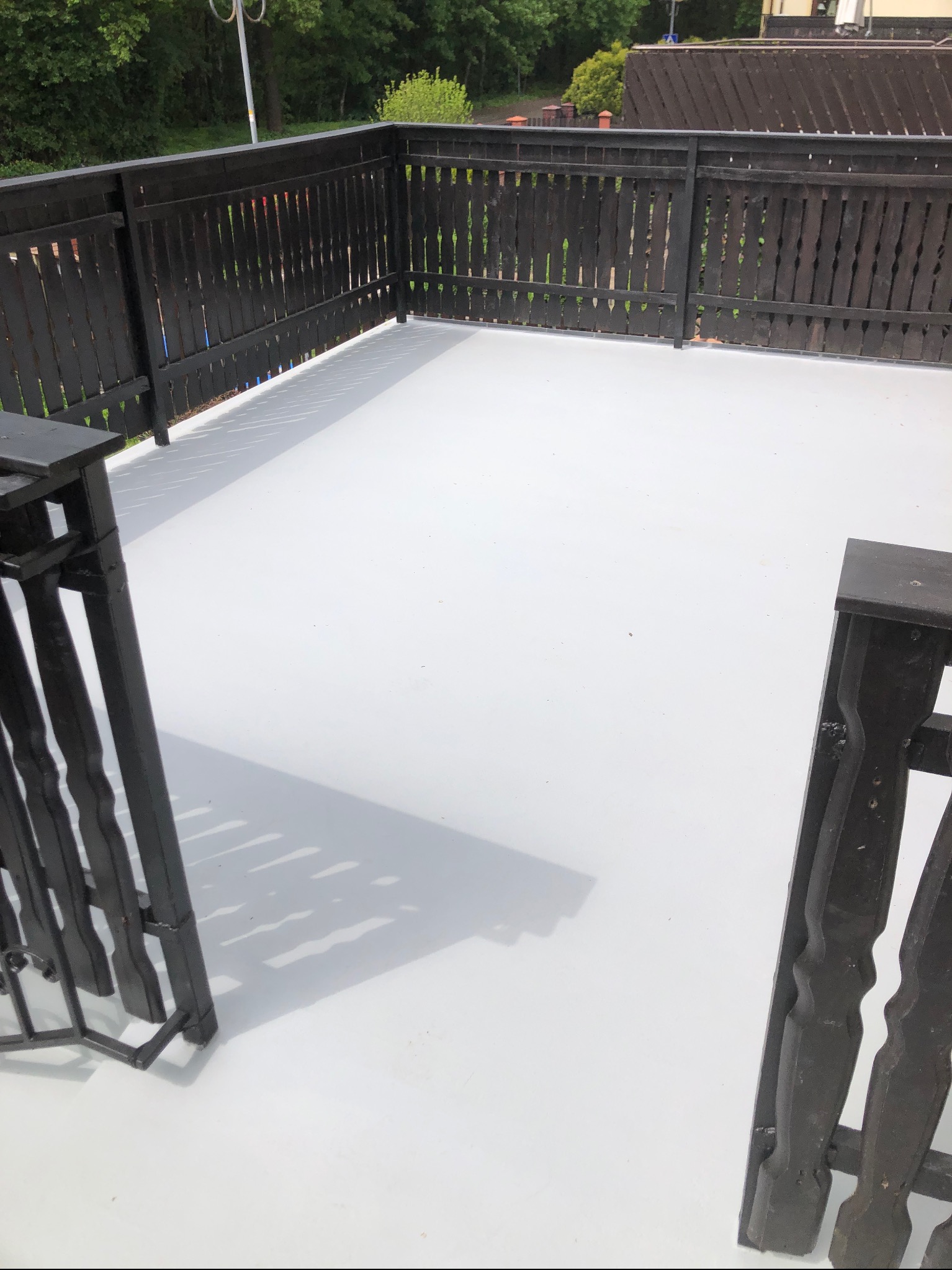 Biała, gładka powierzchnia balkonu z ciemnym, drewnianym ogrodzeniem. Widok z góry, ukazujący odnowioną przestrzeń z wyraźnymi cieniami.