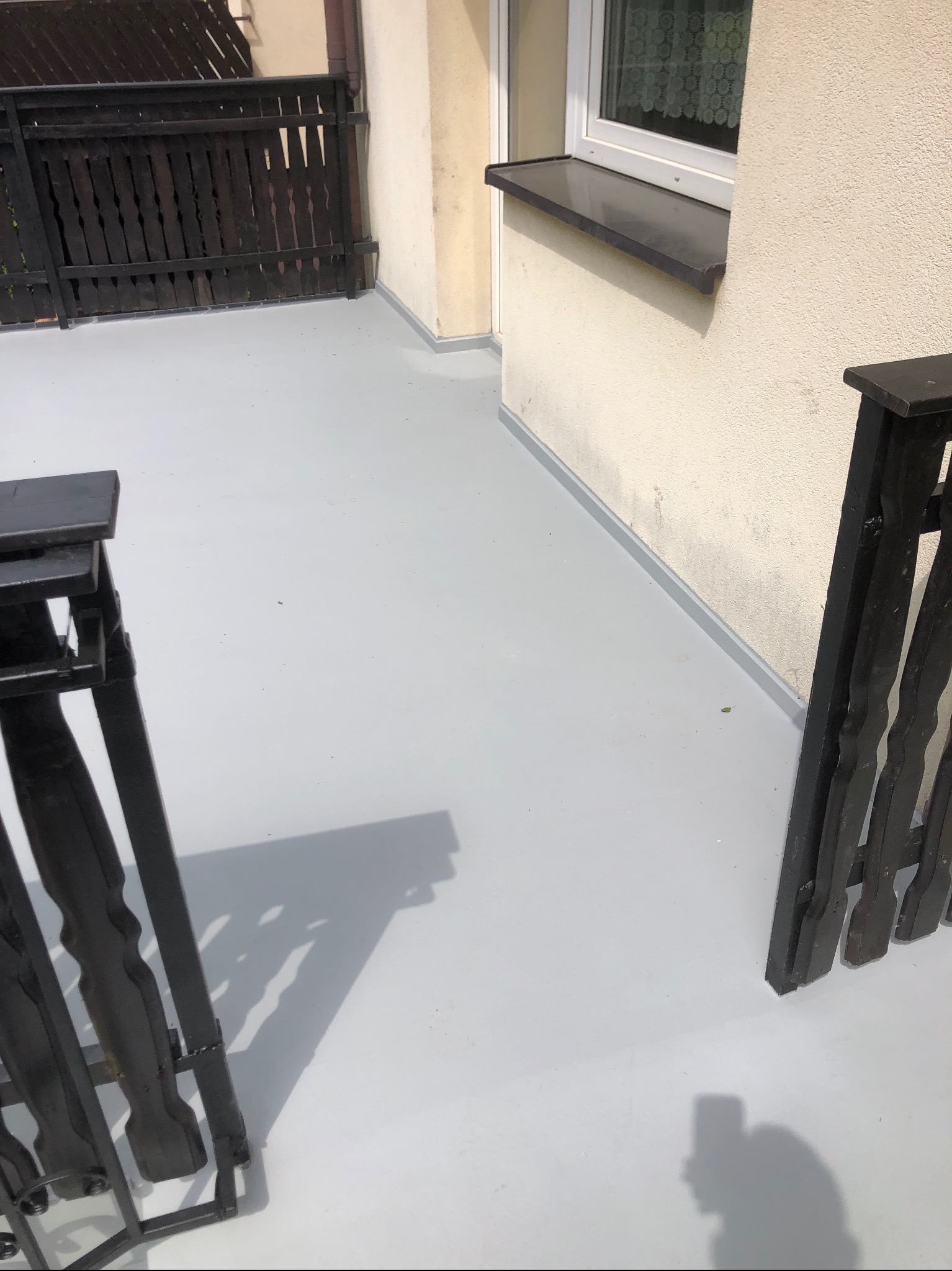 Jasnoszara, gładka powierzchnia balkonu po renowacji, z widoczną drewnianą balustradą i fragmentem elewacji budynku. Minimalistyczny design, czysta przestrzeń.