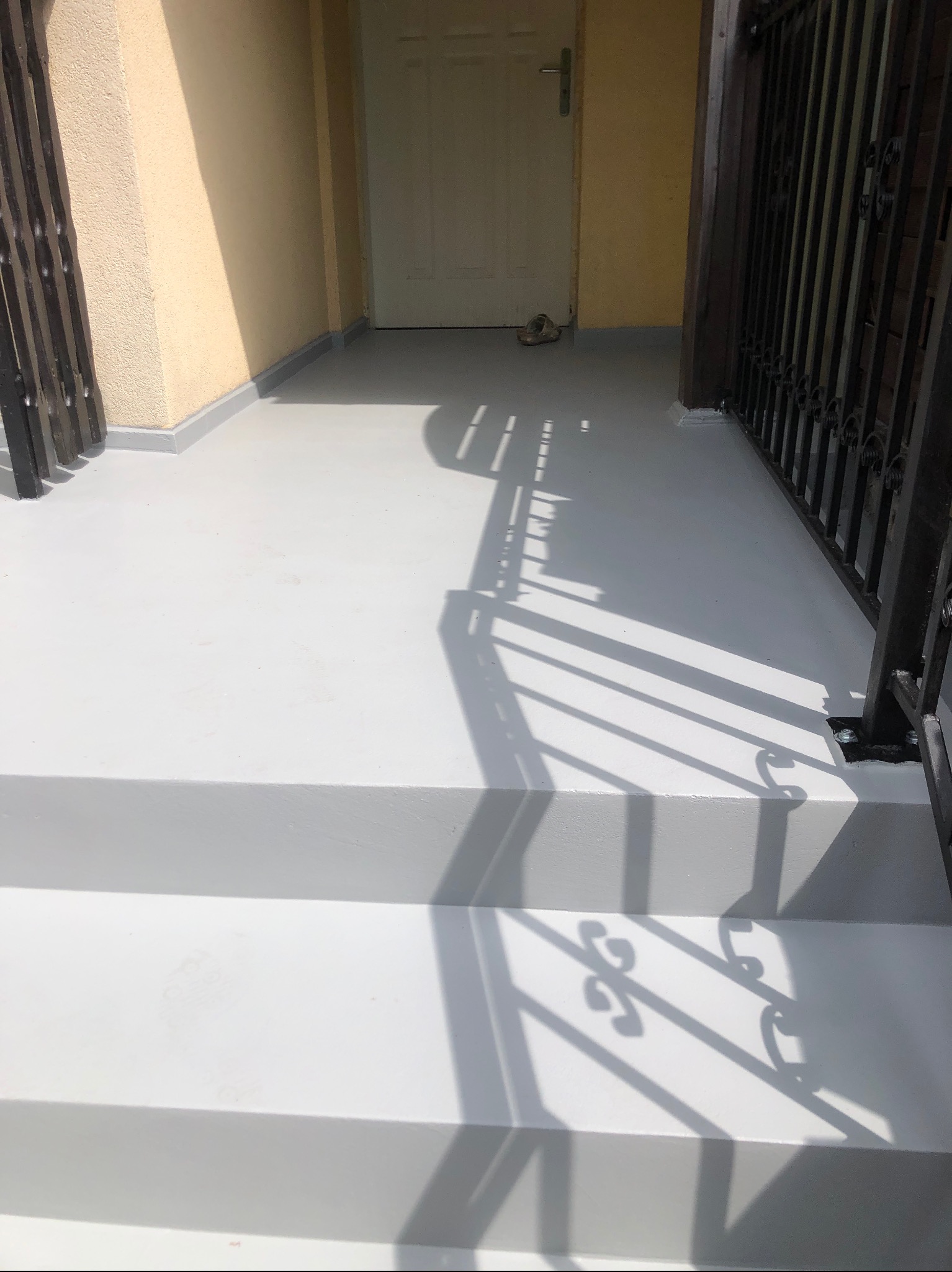 Jasnoszara, gładka powierzchnia balkonu po renowacji, z widocznymi schodami i fragmentem balustrady. Świeże wykończenie, jednolite, matowe. Cień balustrady na posadzce.