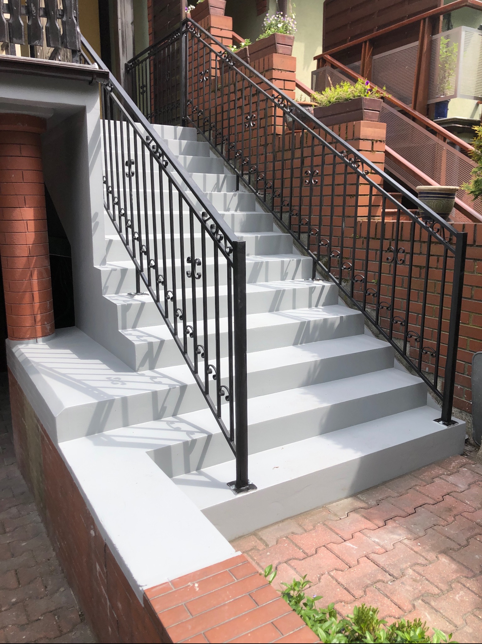 Odnowione schody zewnętrzne z szarą posadzką i czarną, ozdobną balustradą. Widoczne ceglane elementy konstrukcyjne i kostka brukowa.