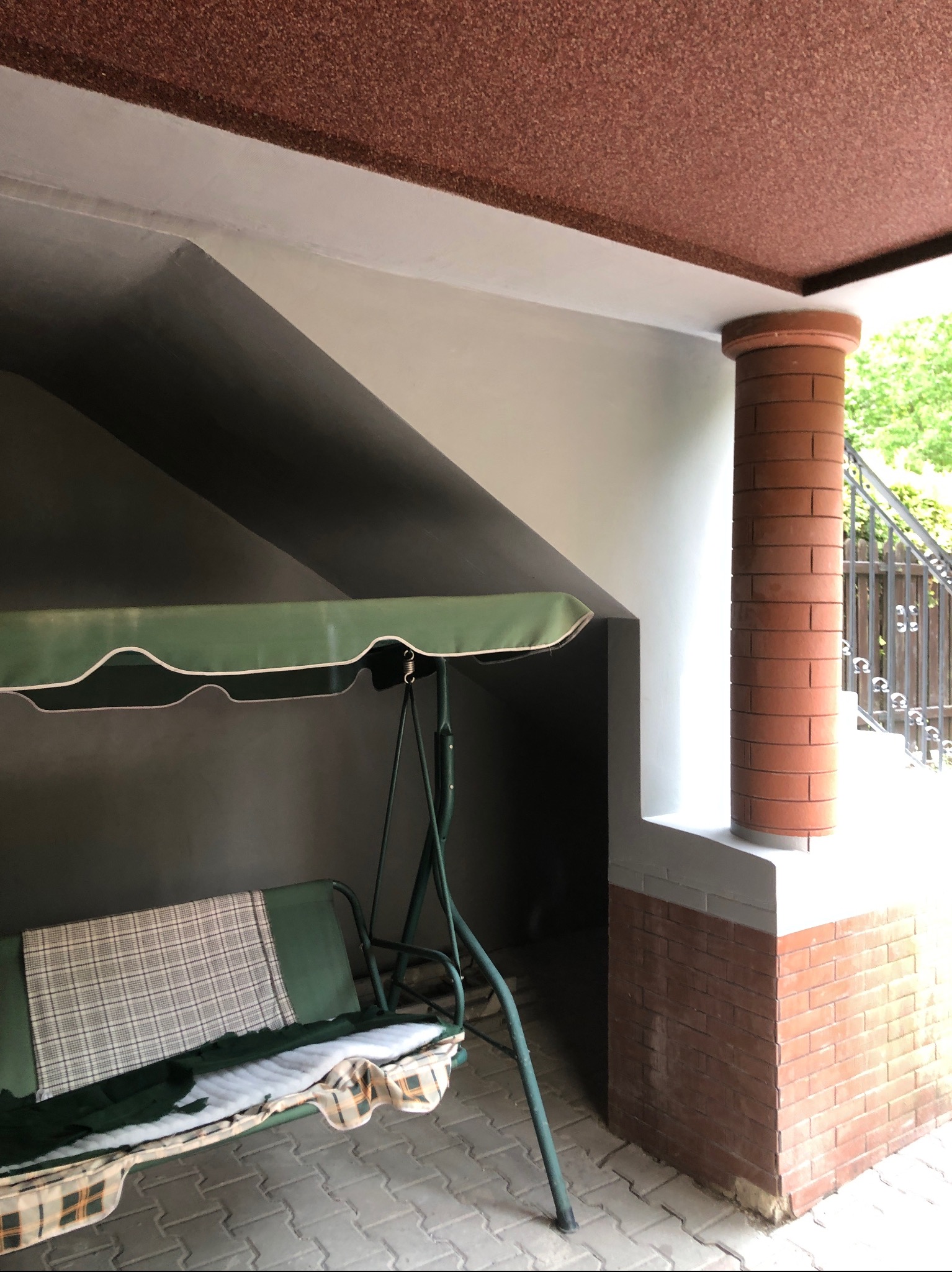 Zadaszony taras z ceglaną kolumną i huśtawką ogrodową. Widoczna kostka brukowa na podłodze i schody z metalową barierką w tle. Ciemne ściany i jasny sufit.