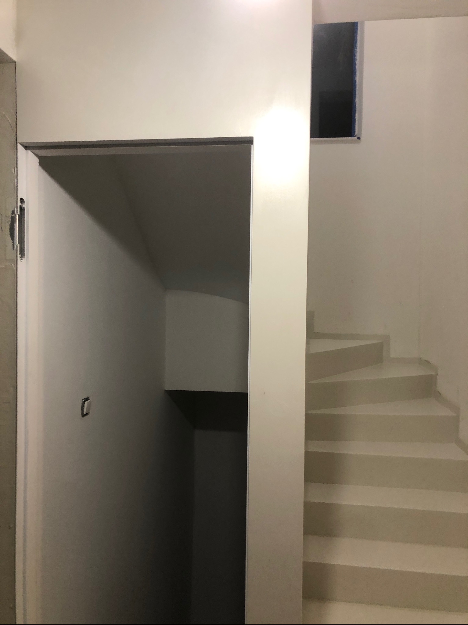 Białe schody prowadzące w górę, obok wejście do ciemnego pomieszczenia. Minimalistyczne wnętrze po remoncie, z oknem w górnej części ściany.