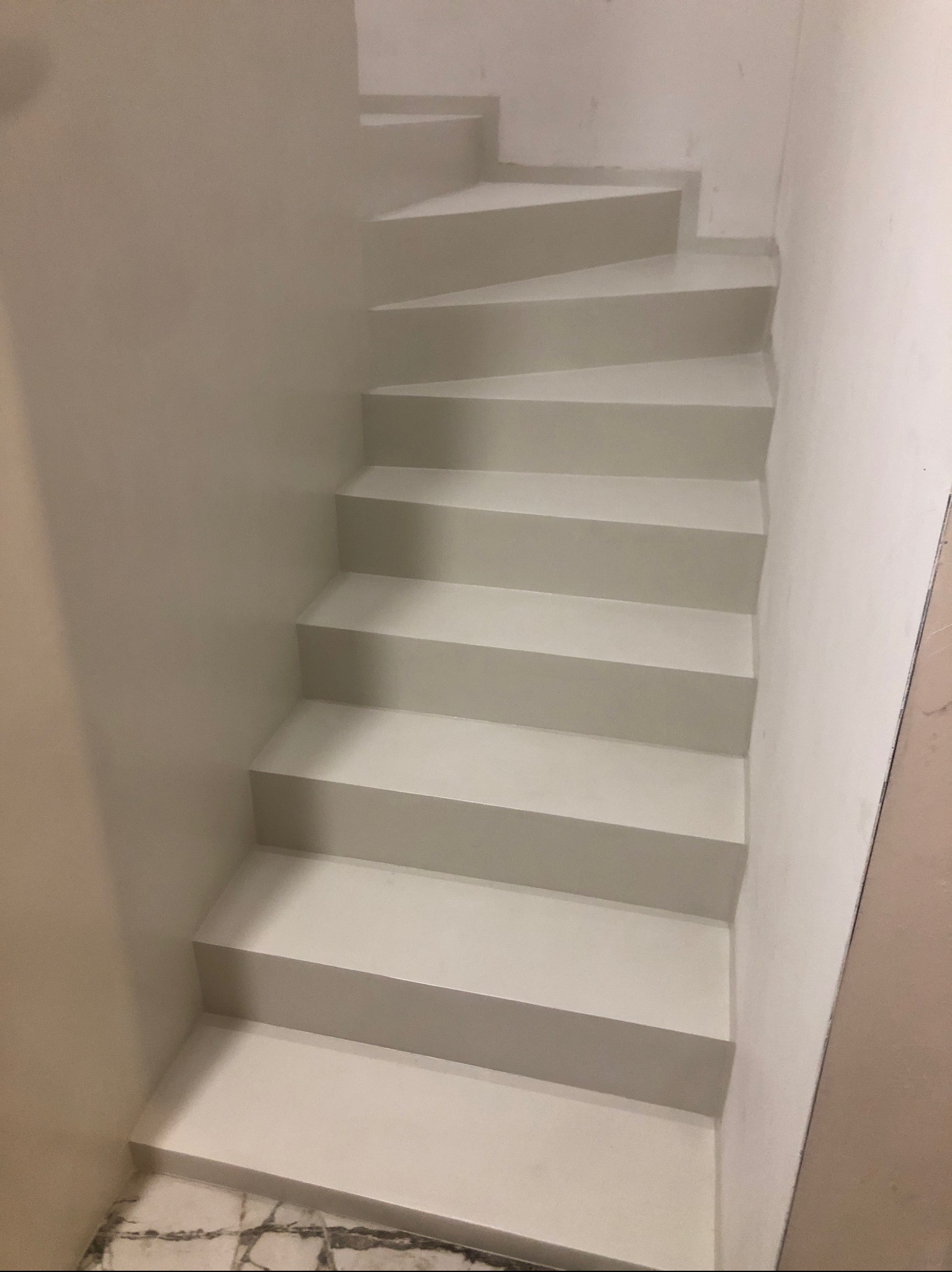 Schody wewnętrzne wykończone jednolitą, jasną powłoką, prawdopodobnie mikrocementem, z widocznym fragmentem marmurowej podłogi u dołu.