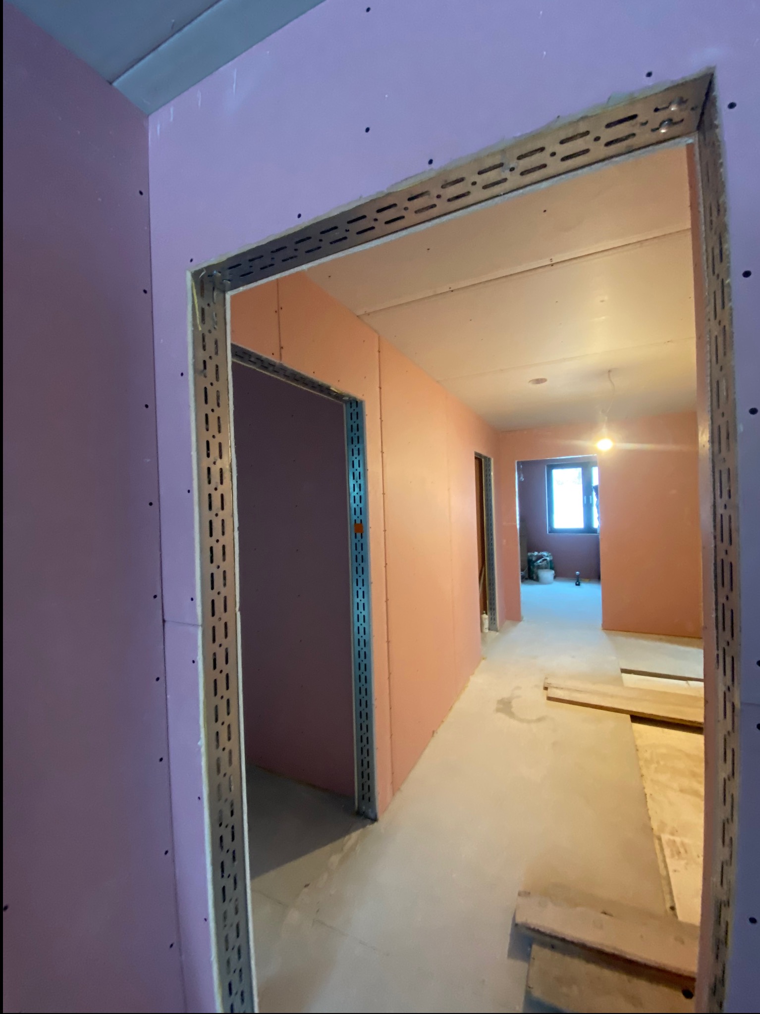 Korytarz w trakcie prac wykończeniowych: ściany obłożone płytami karton-gips w odcieniach różu, metalowe profile przy otworach drzwiowych, surowa podłoga.