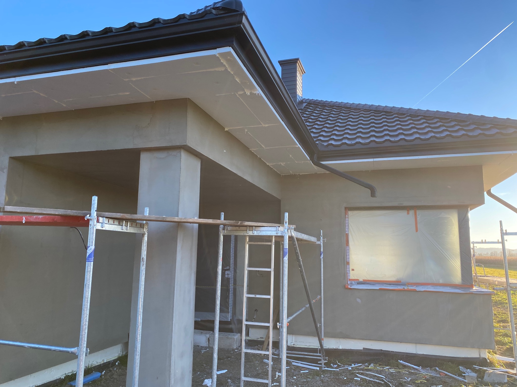 Dom w trakcie prac elewacyjnych: szare ściany, rusztowania, zabezpieczone okno folią. Widoczne detale wykończenia dachu i orynnowania. Słoneczny dzień.