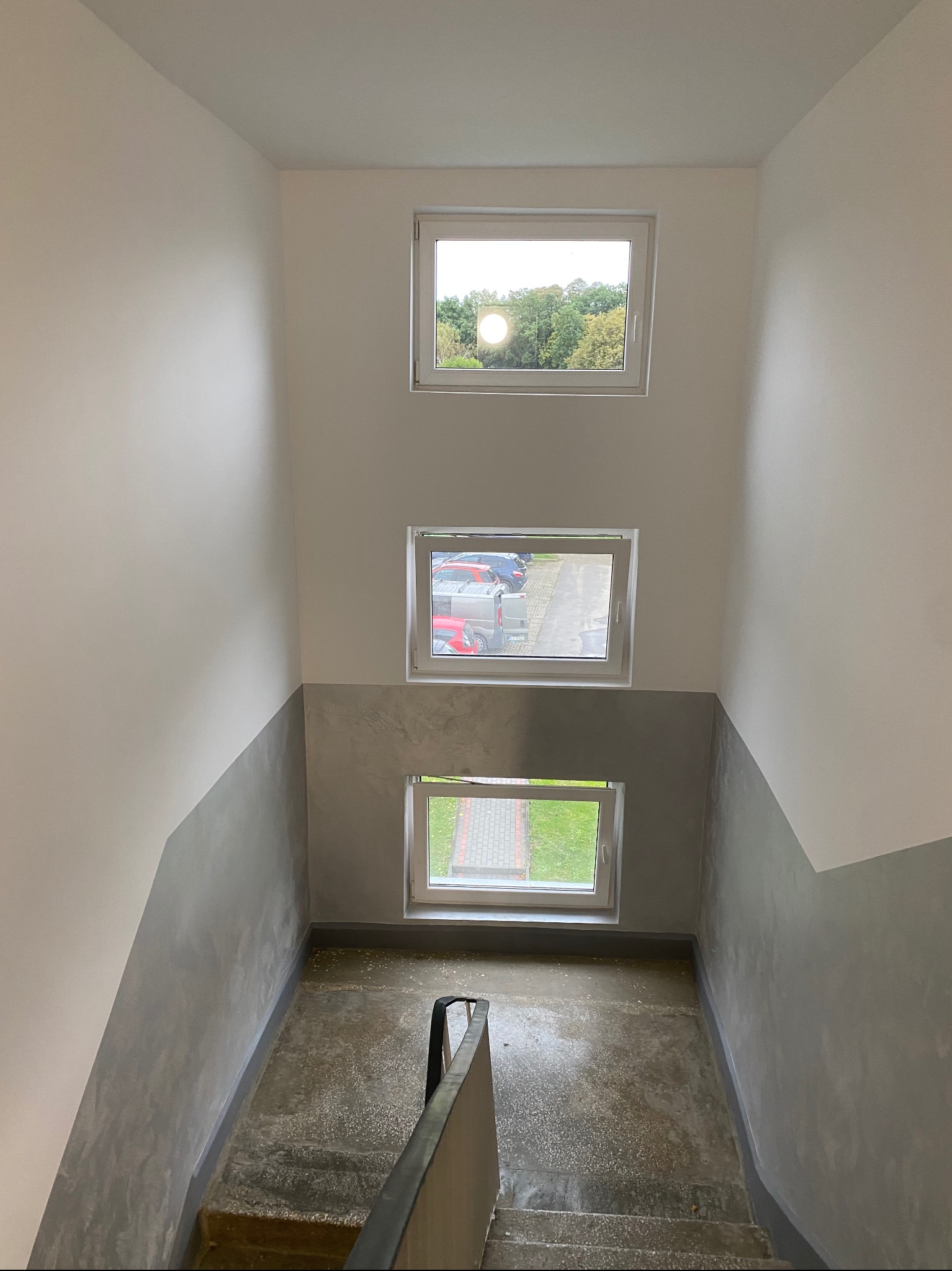 Klatka schodowa po remoncie: białe ściany z szarym pasem na dole, trzy okna na różnych wysokościach wpuszczają światło dzienne. Widoczne schody i fragment poręczy.