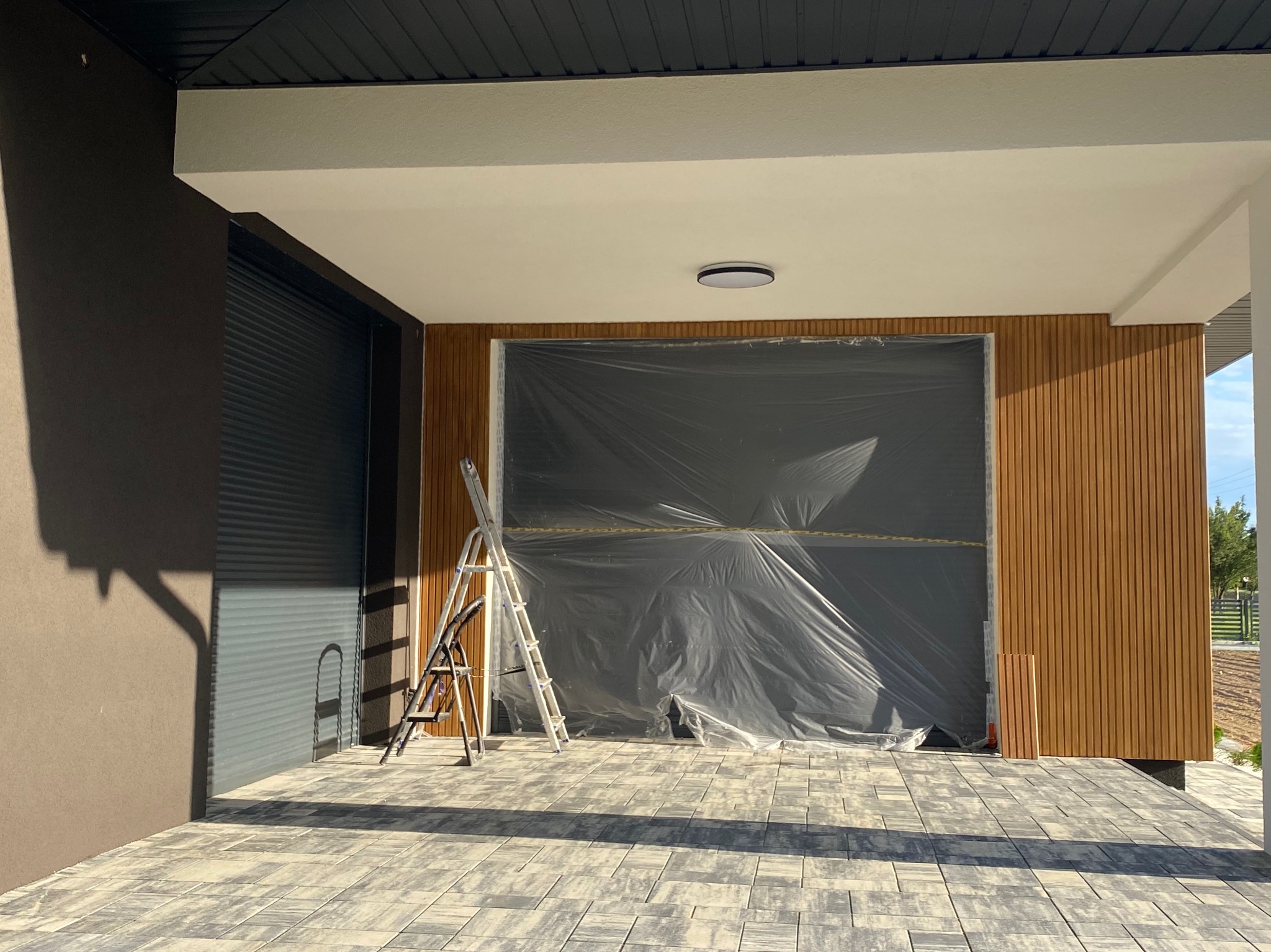 Przygotowania do prac elewacyjnych: zabezpieczona folią ściana z drewnianą okładziną, drabina oparta o ścianę, kostka brukowa na podłożu, cień drzewa na elewacji.