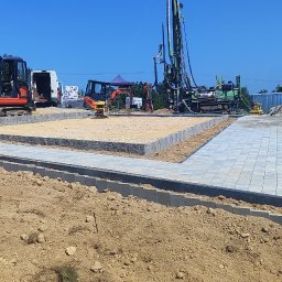 Kamil Góral - Plac budowy z mini koparkami i wiertnicą. Część terenu wyłożona kostką brukową, część przygotowana podłoże. Widoczne materiały budowlane i pracownicy.