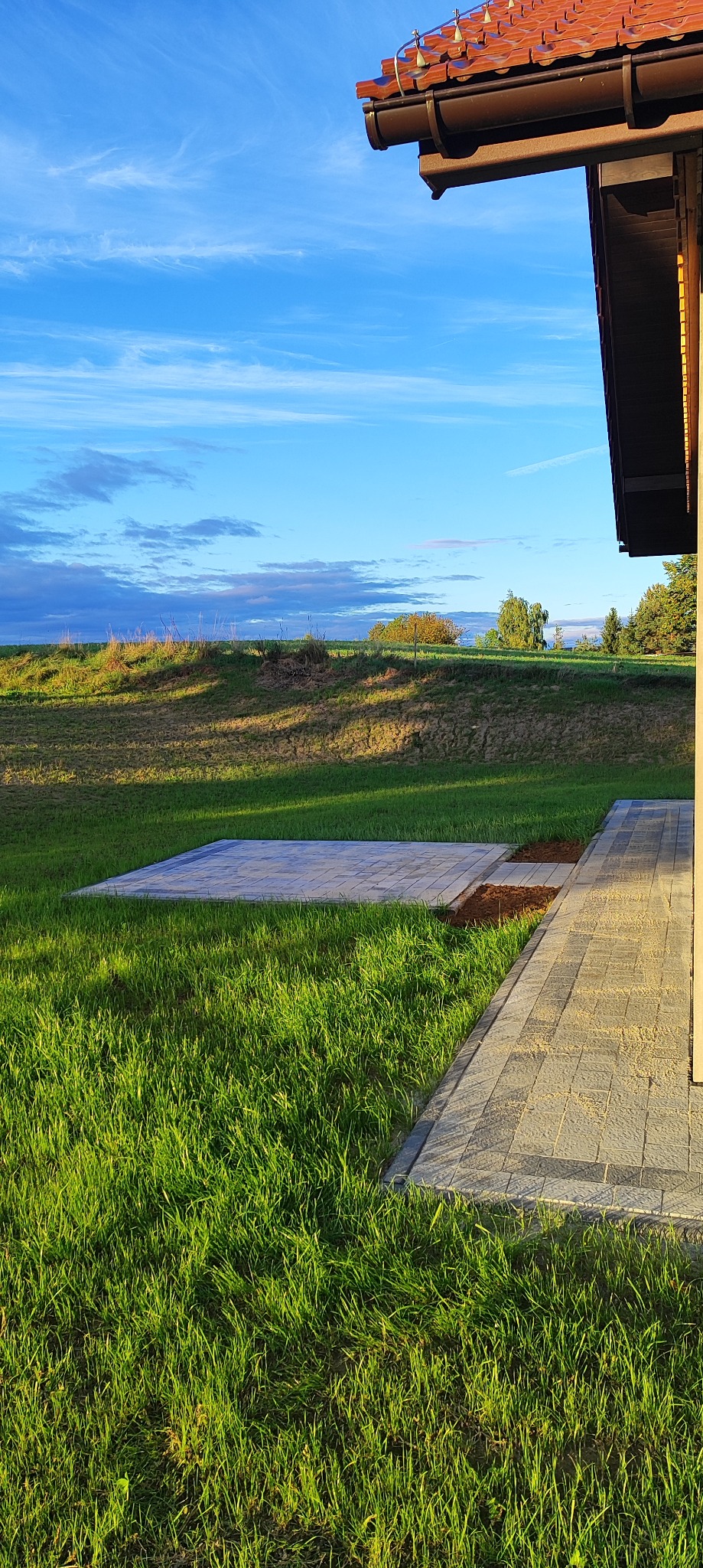 Fragment posesji z ułożoną kostką brukową w trawie, widok z perspektywy trawnika, w tle pagórkowaty krajobraz pod błękitnym niebem. Część dachu budynku w prawym górnym rogu.