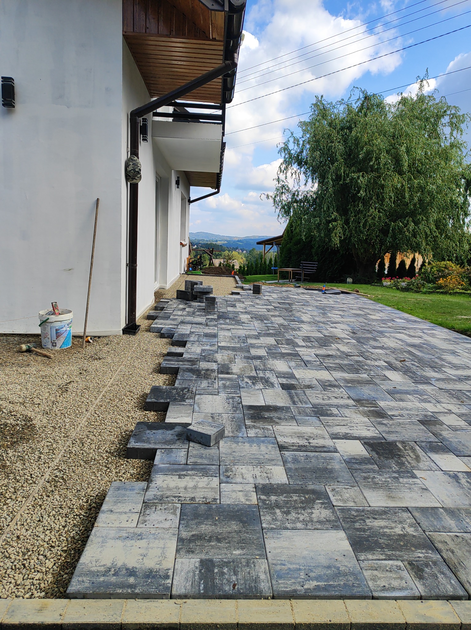 Fragment układania szarej kostki brukowej na posesji, widoczne narzędzia i przygotowane podłoże. W tle budynek z białymi ścianami i zielony ogród.