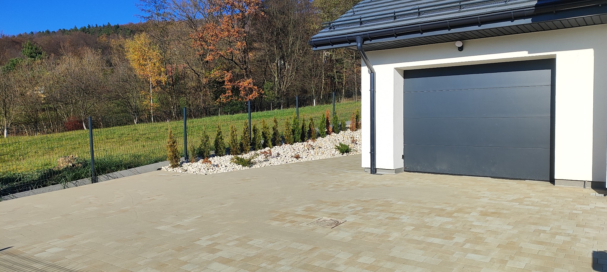 Jasna kostka brukowa przed garażem z szarą bramą, obok ozdobny pas z kamieni i tuj oraz ogrodzenie panelowe na tle zieleni i drzew w jesiennych barwach.