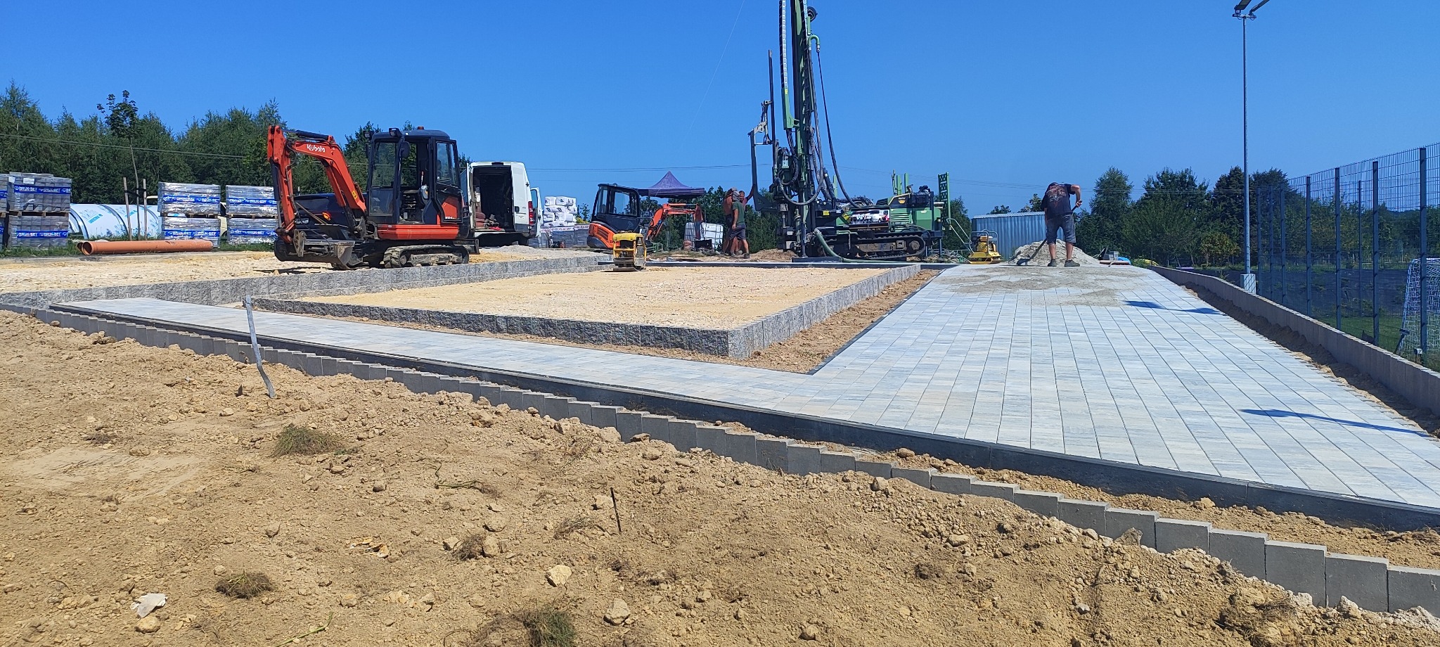Plac budowy z mini koparkami i wiertnicą. Część terenu wyłożona kostką brukową, część przygotowana podłoże. Widoczne materiały budowlane i pracownicy.