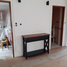 Andrzej Staszowski - Hall z konsolą w stylu klasycznym: drewniany blat, czarna podstawa. Na ścianie dekoracyjna sztukateria i dwa czarne kinkiety. Widoczne przygotowania do układania parkietu.