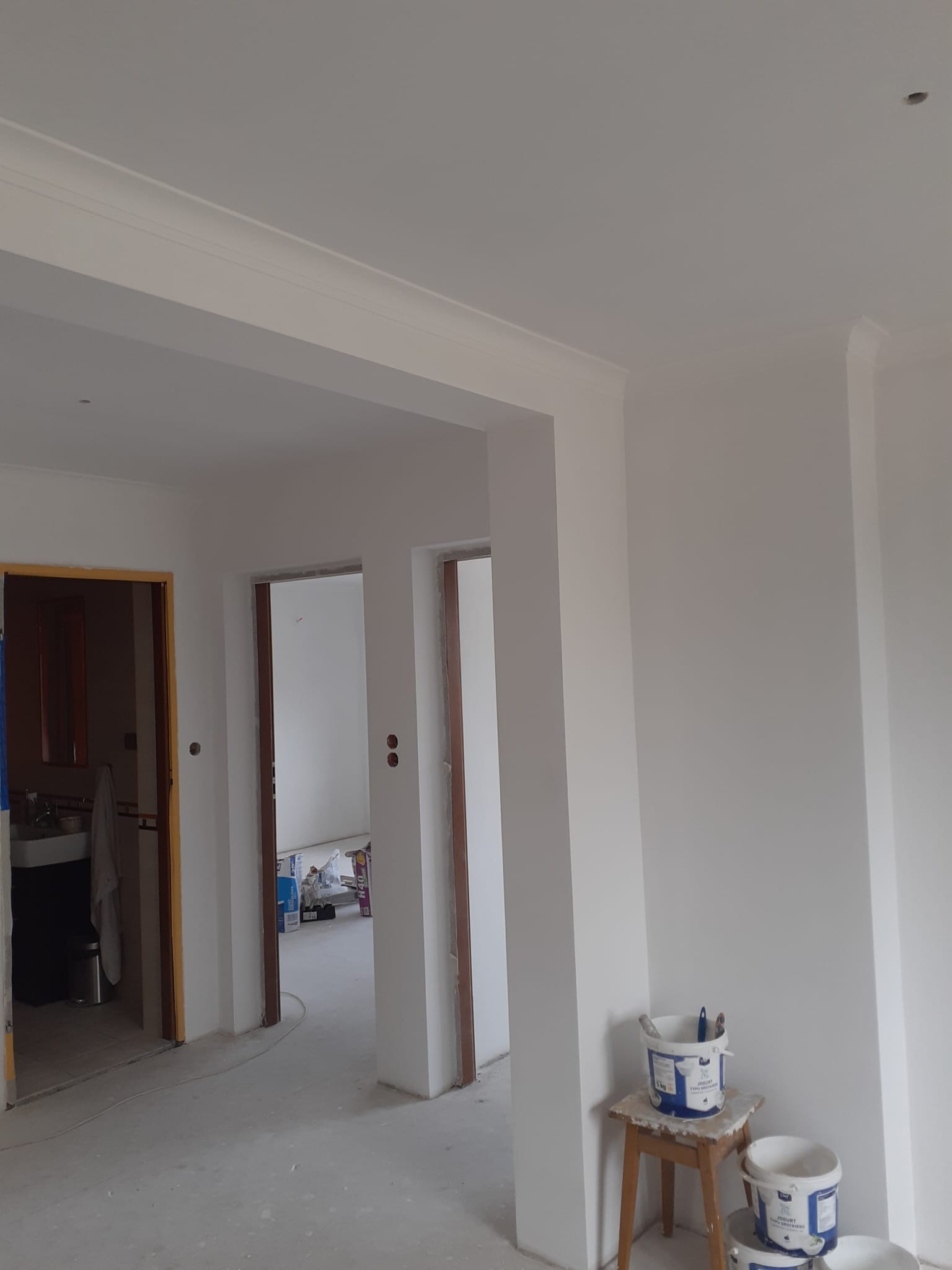 Białe ściany po malowaniu w jasnym wnętrzu z trzema otworami drzwiowymi. Widoczne wiaderka z farbą i narzędzia malarskie na stołku. Listwy dekoracyjne przy suficie.