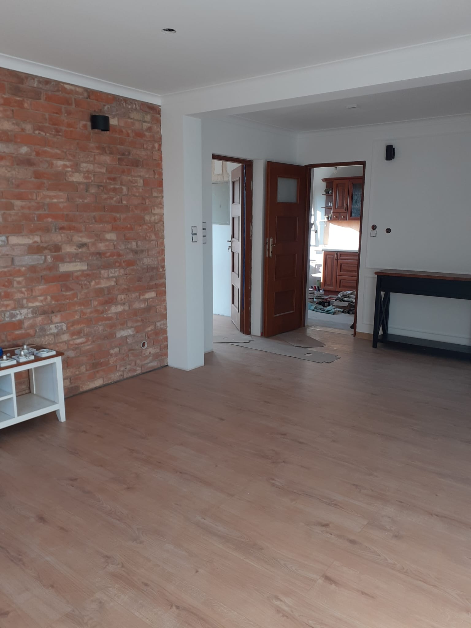 Jasne panele podłogowe w pokoju z ceglaną ścianą i białymi listwami. Widoczne drzwi do kuchni w trakcie remontu. Minimalistyczny stolik z boku.