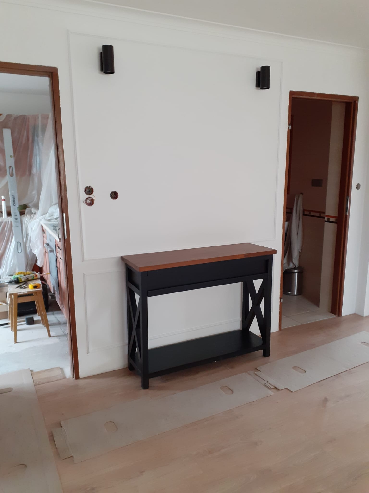 Hall z konsolą w stylu klasycznym: drewniany blat, czarna podstawa. Na ścianie dekoracyjna sztukateria i dwa czarne kinkiety. Widoczne przygotowania do układania parkietu.