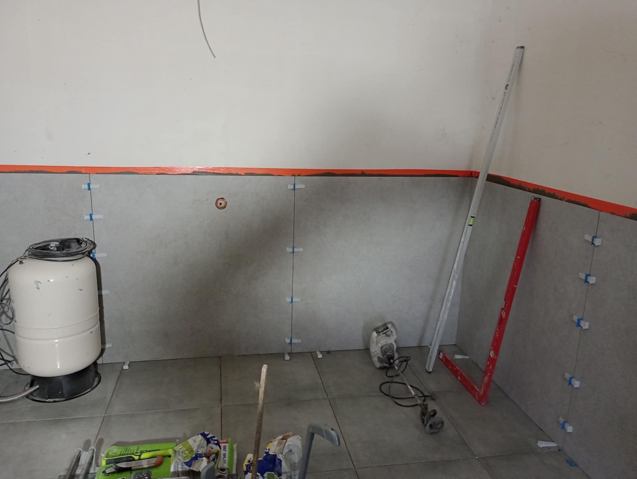 Ściana w trakcie układania szarych płytek z użyciem poziomicy i krzyżyków dystansowych. Widoczny zbiornik hydroforowy i narzędzia glazurnicze.